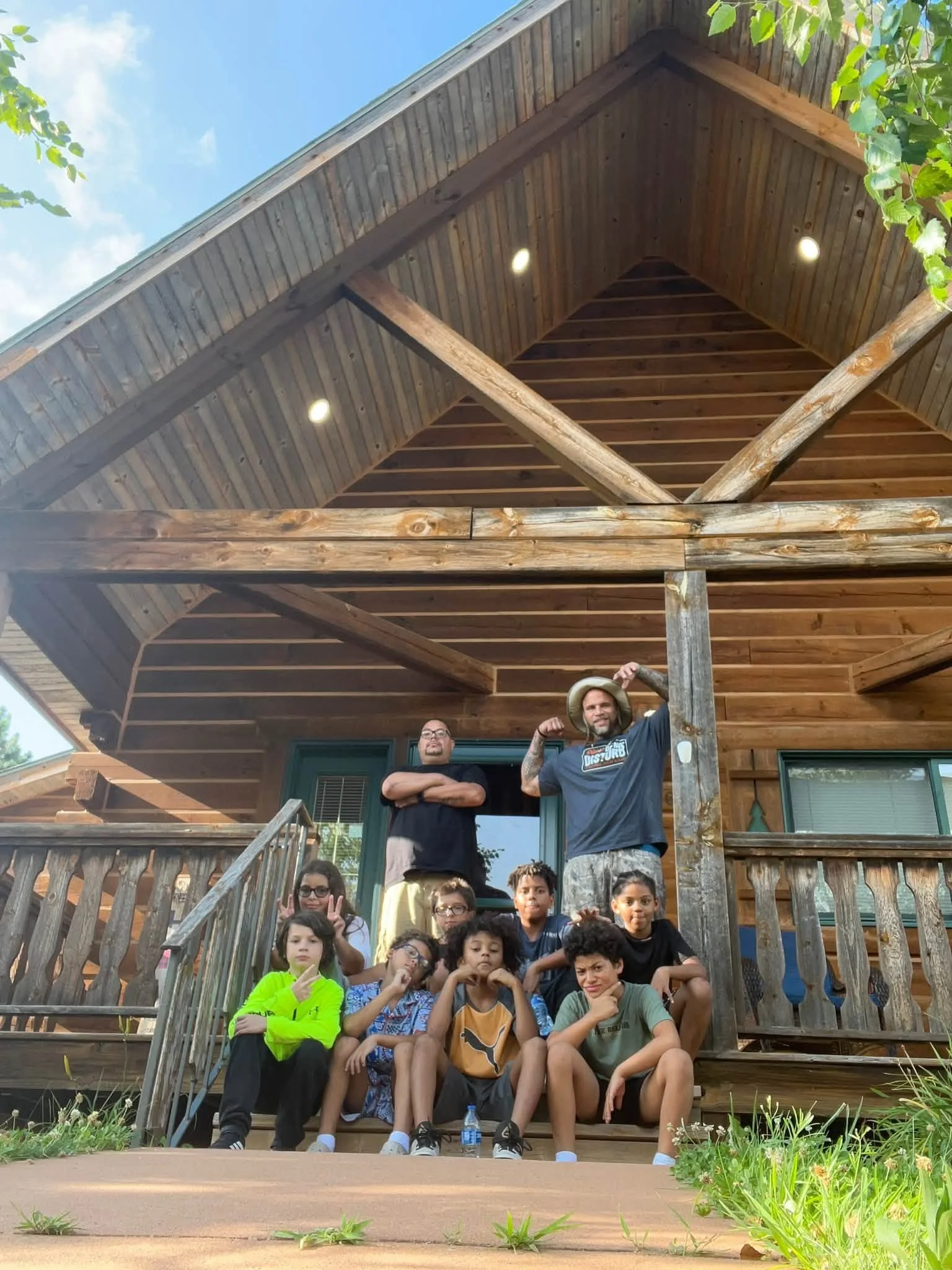 A group of children and two adults posing on the steps of a wooden house with a large group of children sitting in front of them, some making peace signs or other hand gestures, in a sunny outdoor setting.