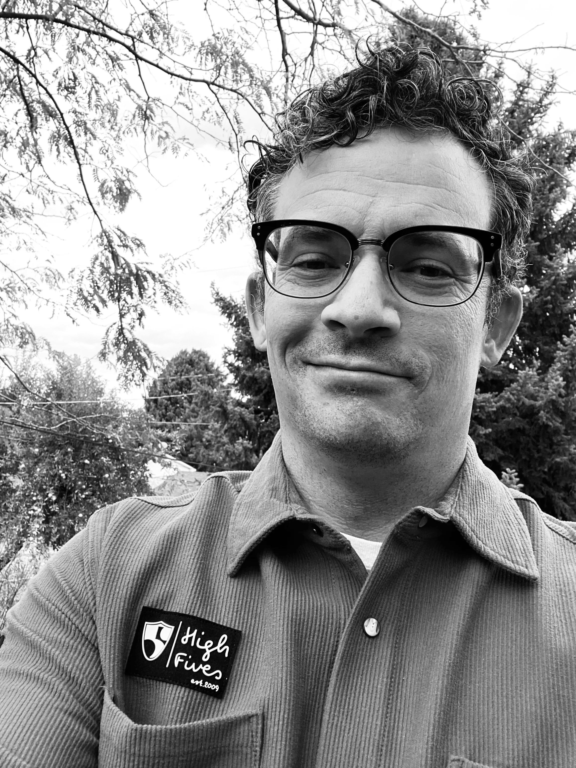 Shane Farver has a closed-mouth smile in front of some trees. He has curly black hair that is parted off to one side and browline glasses. He is wearing a corduroy shirt  with a High Fives Foundation logo. He has a killer mustache (not pictured). 