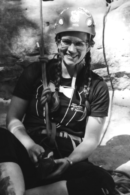Miranda Scott smiles while wearing a helmet and being tied into a rope. She has glasses and a "tuxedo shirt" from ACF on. Her hair is in two braids. 