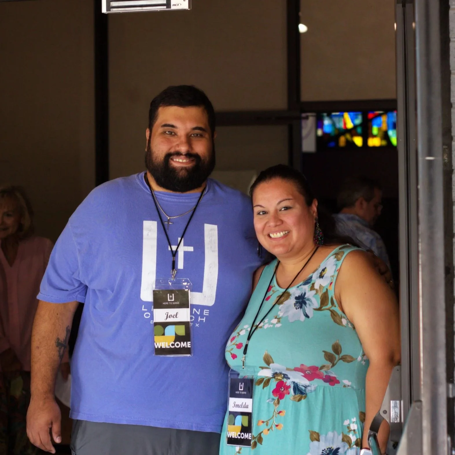 A man and woman smiling at a welcoming event. The man is wearing a blue T-shirt and the woman is in a floral dress. Both have name tags around their necks.
