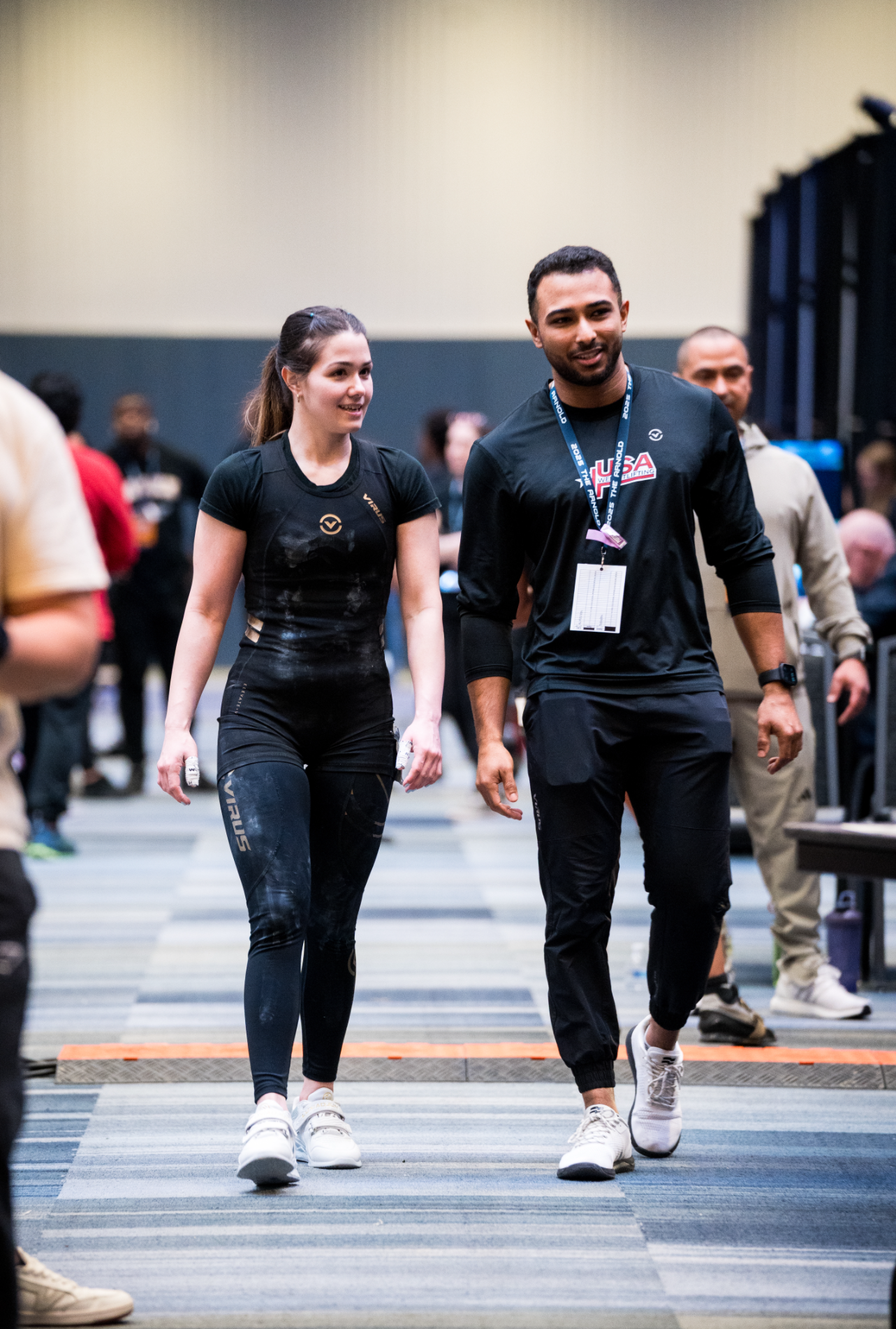 Two athletic people walking together in a gym or sports facility; a woman in black sportswear and a man in black athletic clothing with a badge, both smiling.