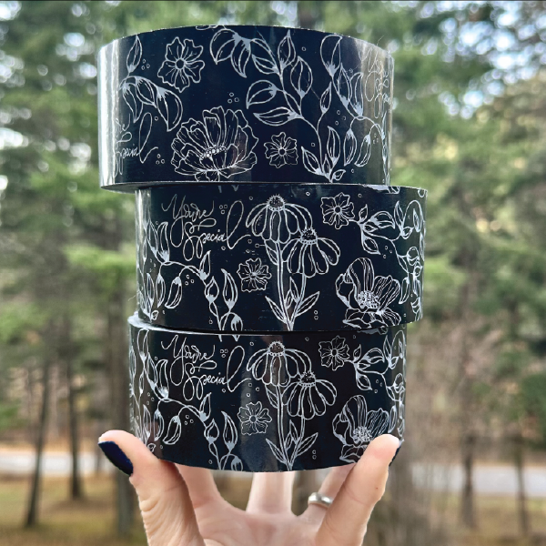 Three stacked rolls of decorative black packing tape with white floral designs and the text "You're special" held by a hand against a blurred forest background.