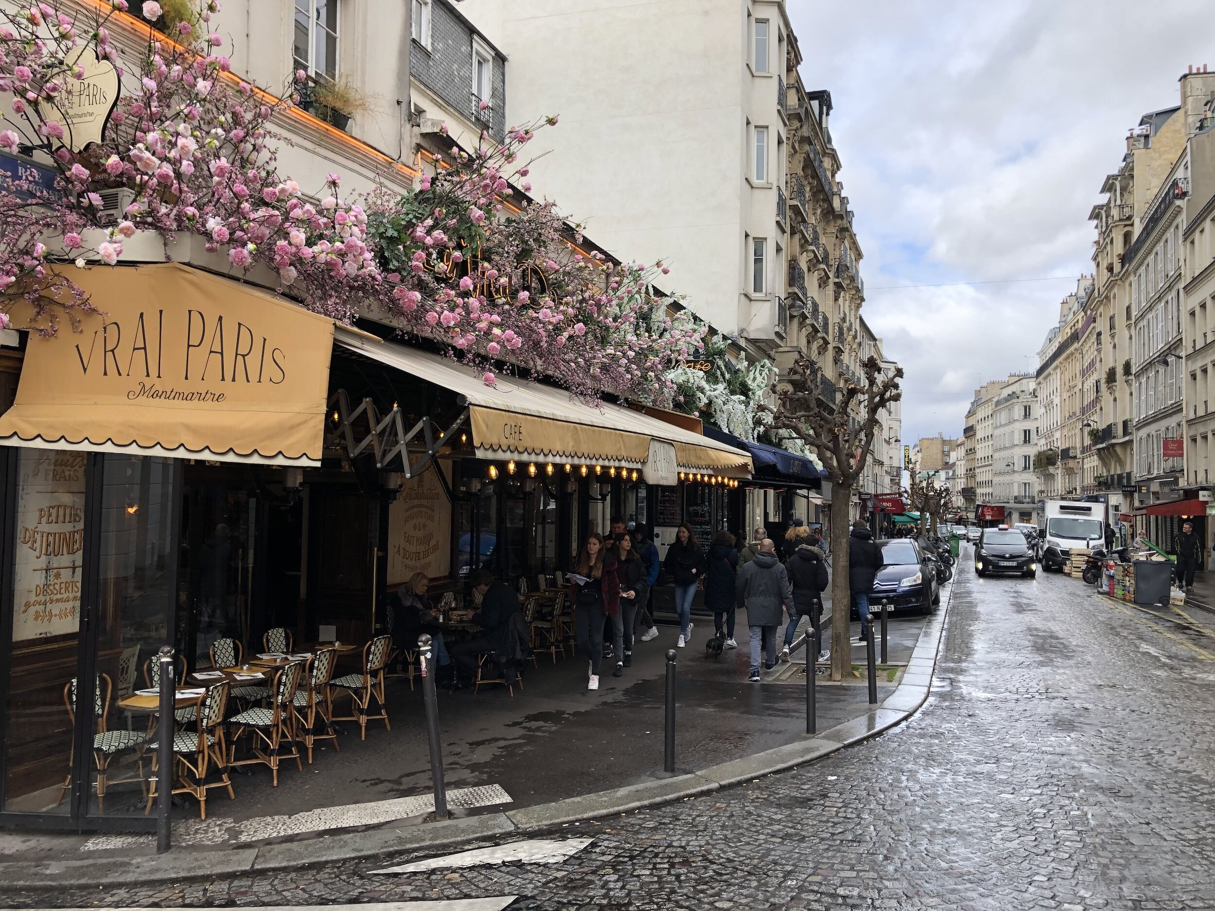 Street scene in Montmartre, Paris, featuring Vrai Paris café with outdoor seating and floral decorations, people walking on the cobblestone sidewalk, and parked cars along the street.