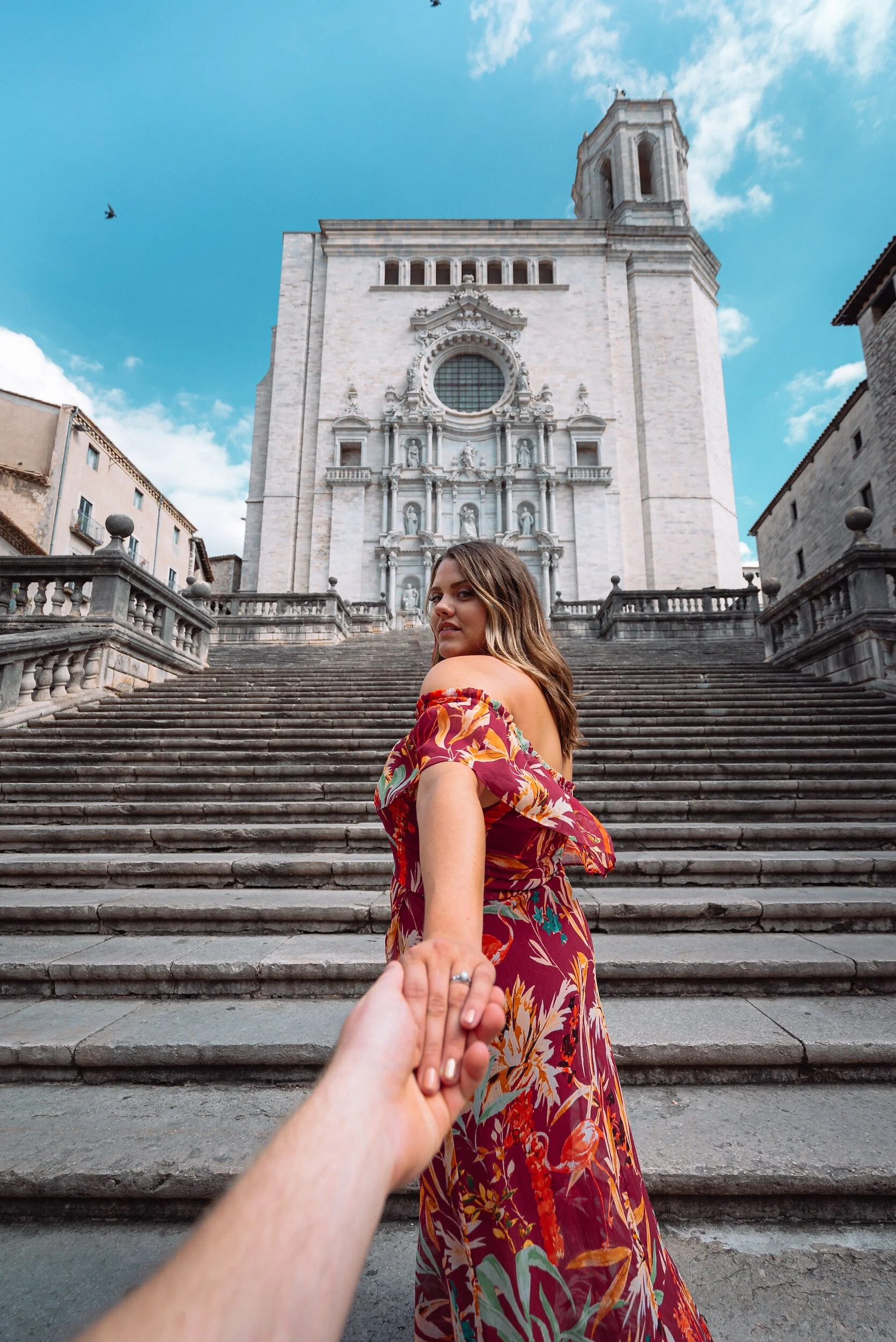 Woman in floral dress on ancient stone steps, holding a hand, with historic building in background, blue sky.
