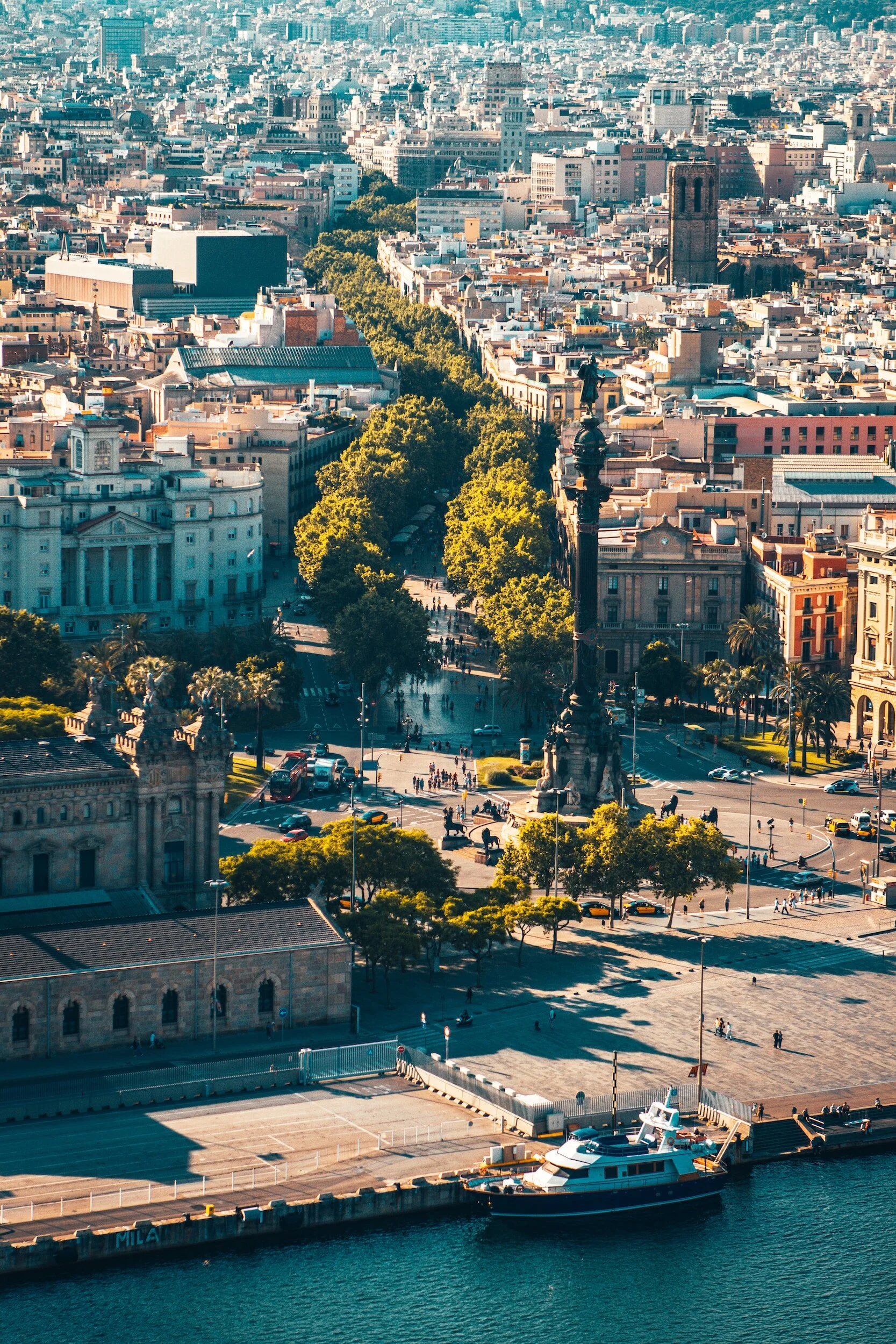 Aerial view of Barcelona with the Columbus Monument, surrounded by city buildings and tree-lined streets, near the waterfront with a docked boat.