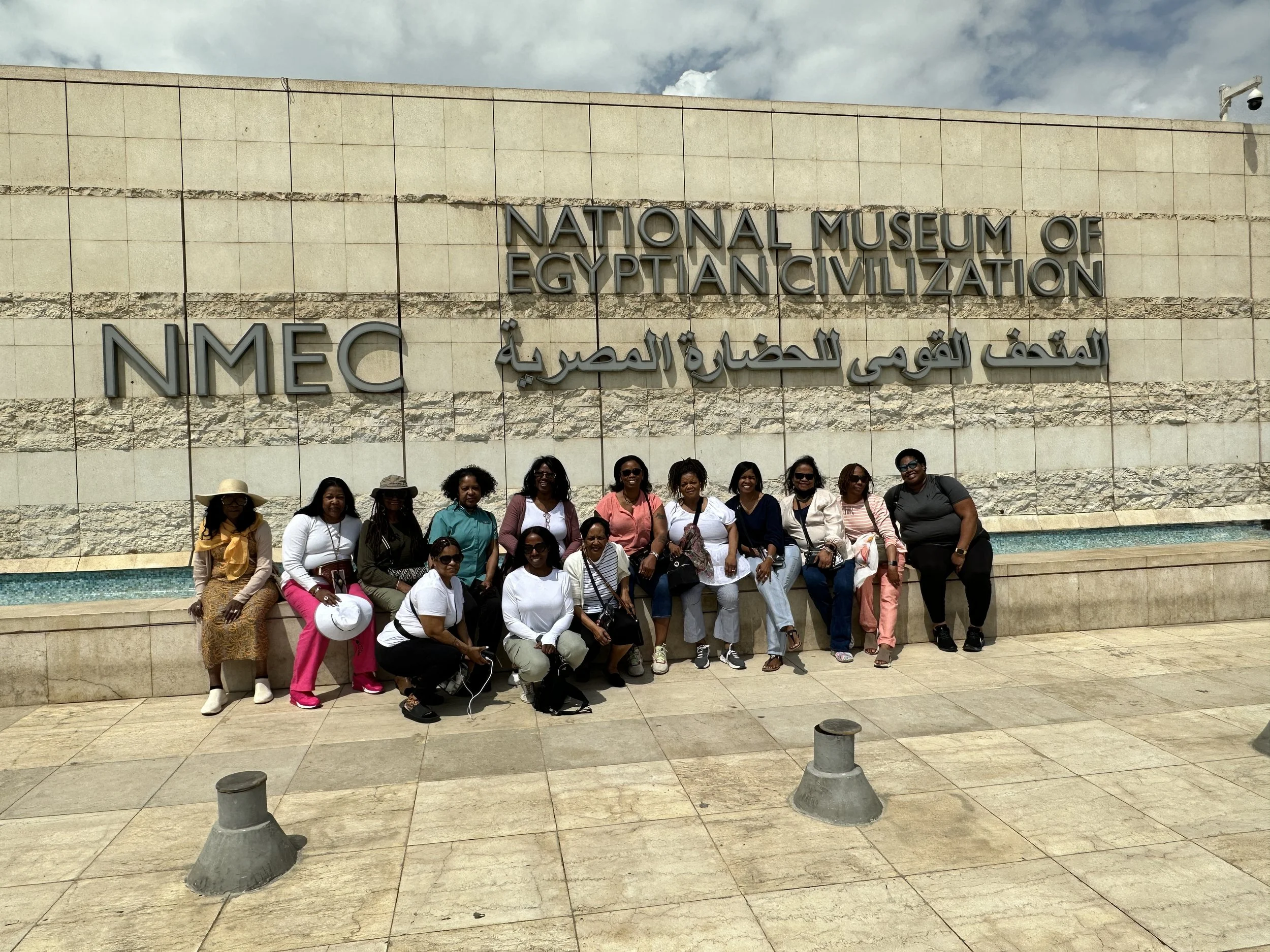 A group of women standing and sitting in front of the sign for the National Museum of Egyptian Civilization (NMC) in Egypt, with some wearing sunglasses and hats, and a mostly cloudy sky above.