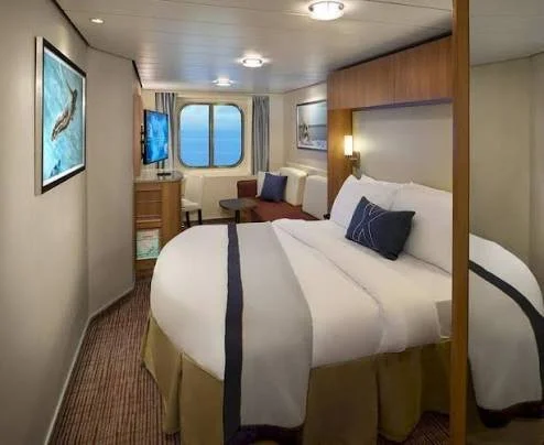 Interior of a cruise ship cabin with a large bed, a window with a view, a small sitting area, and wall art.