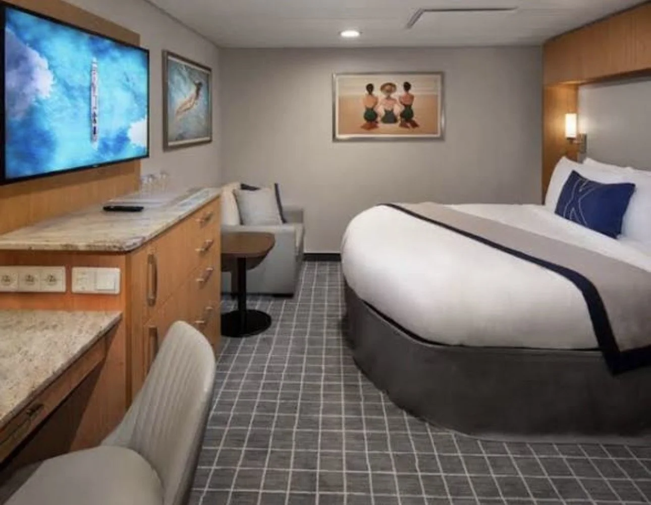 Hotel room with a large bed, TV on a wooden dresser, framed artwork on the walls, sitting area with cushions, and a checkered carpet.
