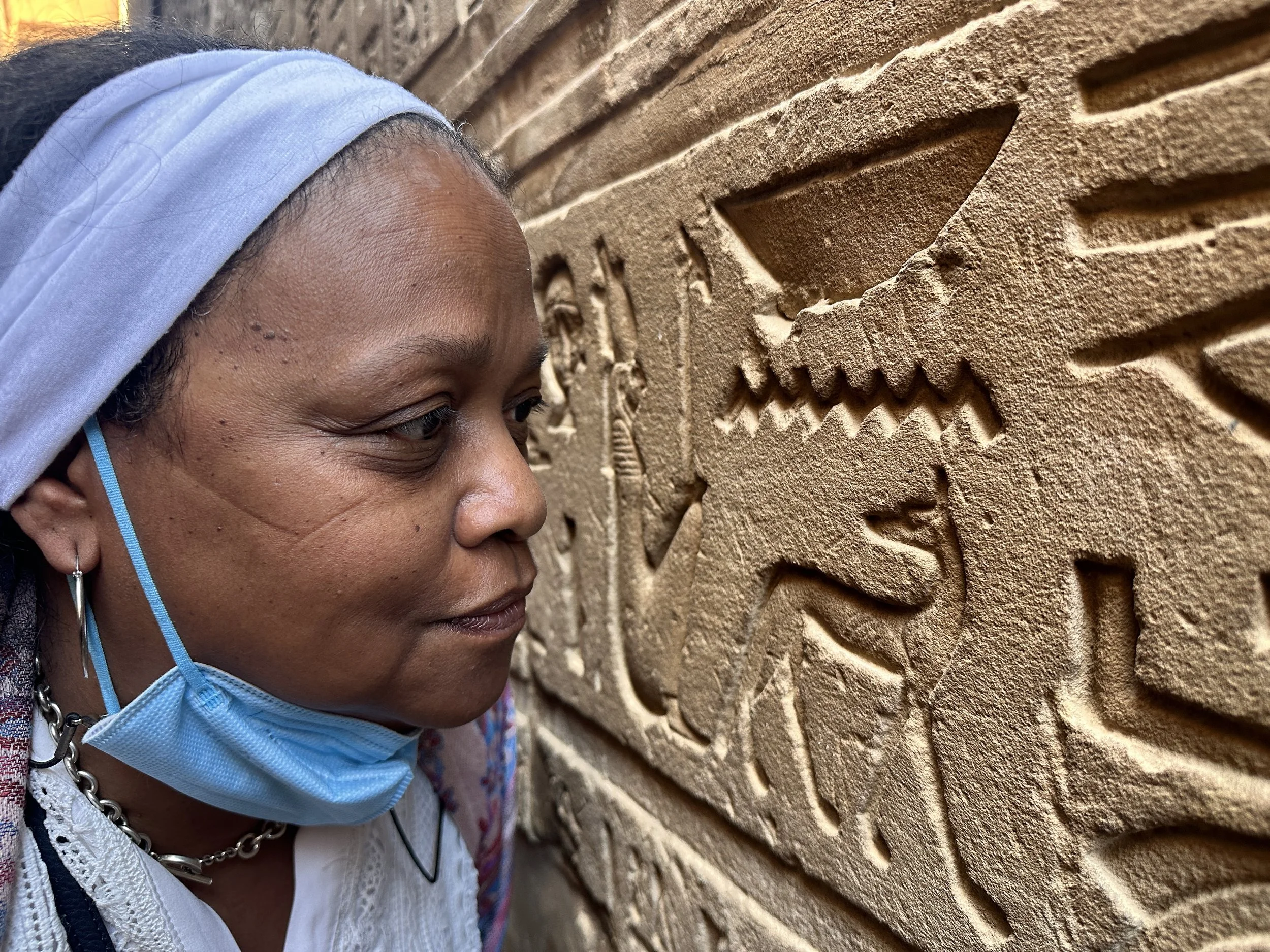 A woman wearing a face mask and headscarf closely studies ancient Egyptian hieroglyphs carved into a stone wall.