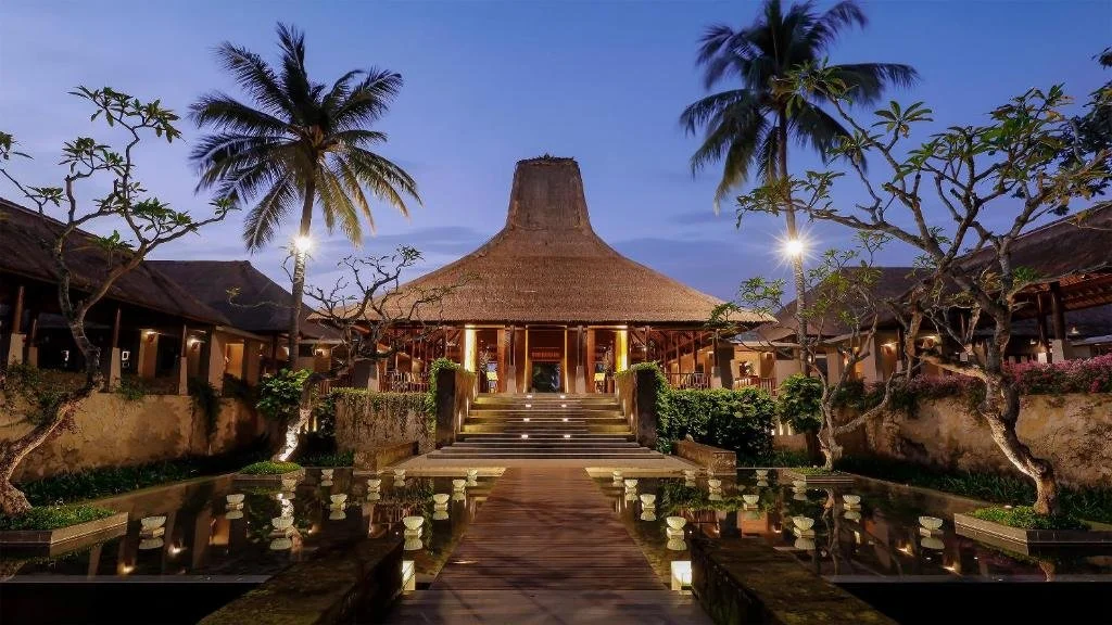 Tropical resort with thatched roof building, palm trees, stone steps, and water features at dusk.