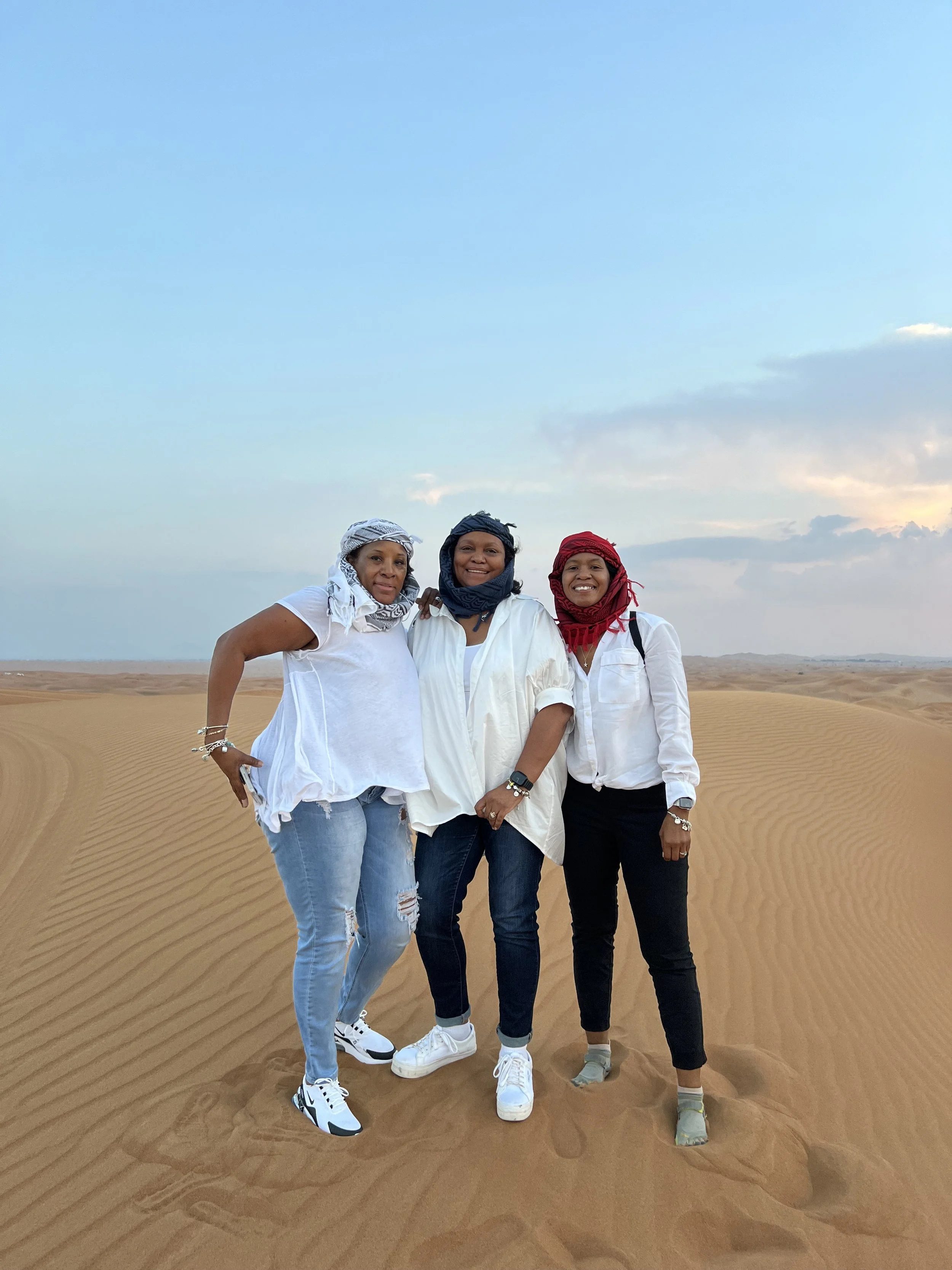 Three women standing in the desert with sand dunes and a partly cloudy sky in the background.