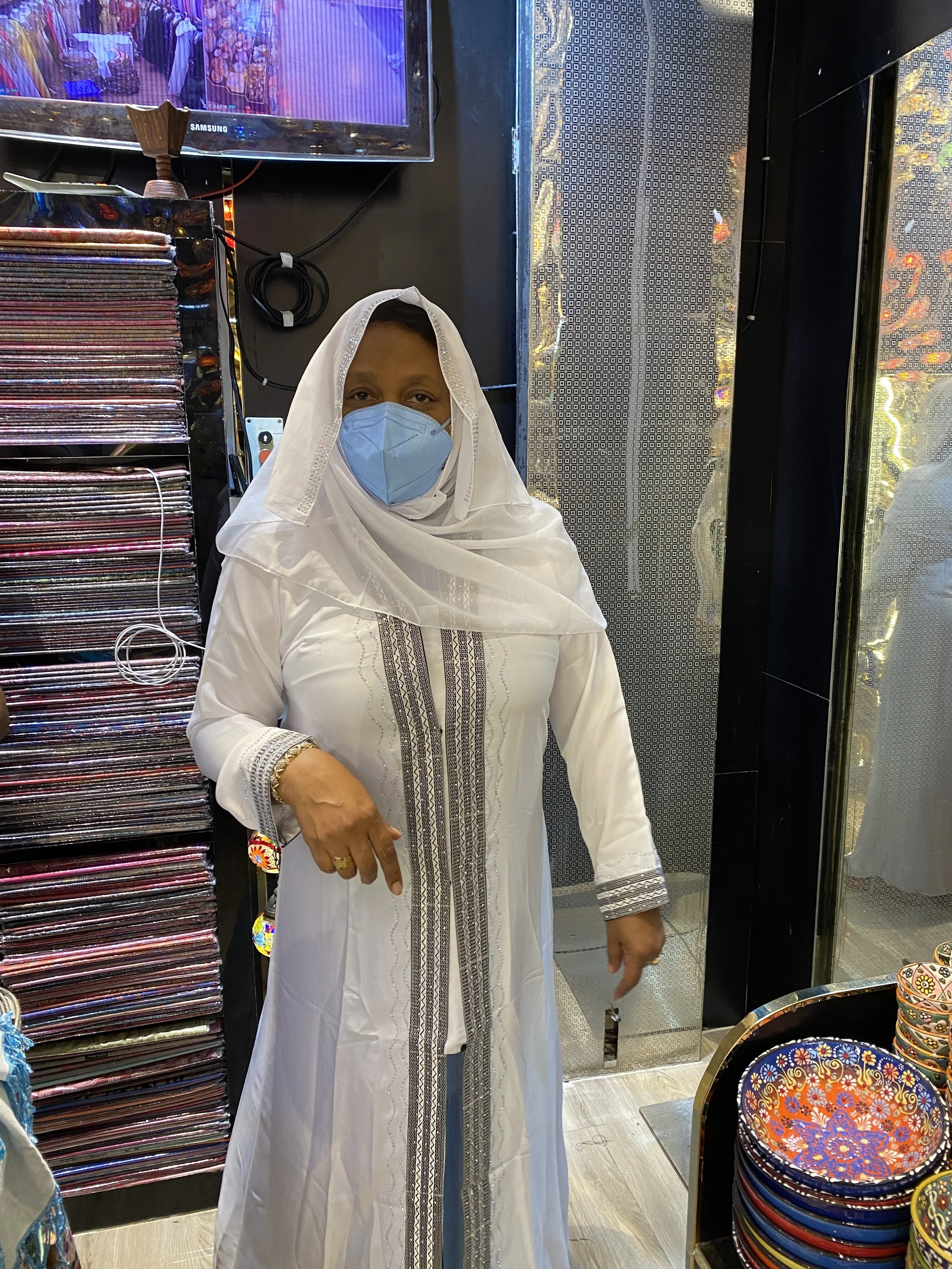 Woman in white traditional clothing and blue face mask standing inside a shop with colorful plates and a rack of fabrics, with a television mounted on the wall behind her.