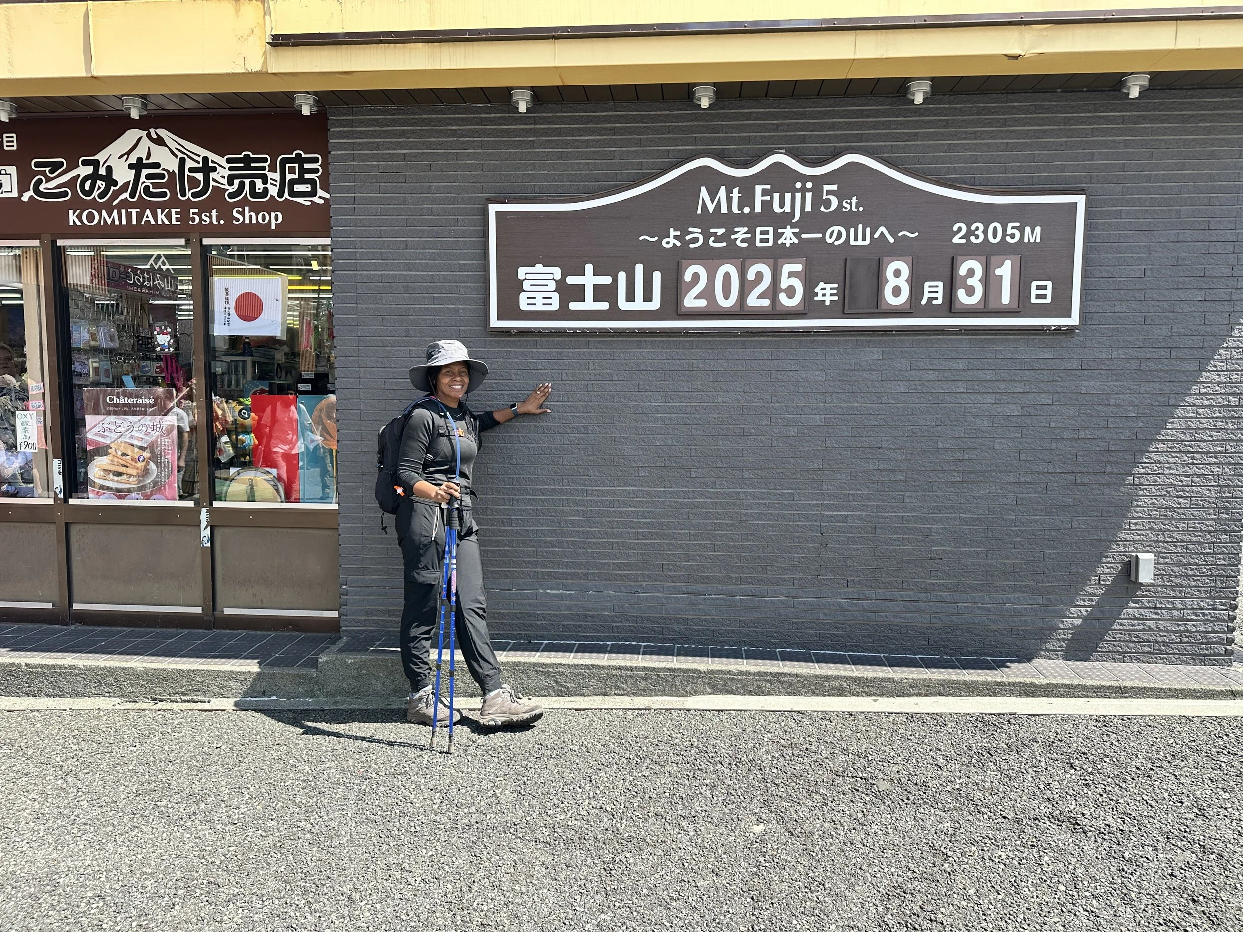 A person in outdoor hiking gear, standing and smiling in front of a sign that reads 'Mt. Fuji 5th Station 2305M' with the date August 31, 2025, on a sunny day.