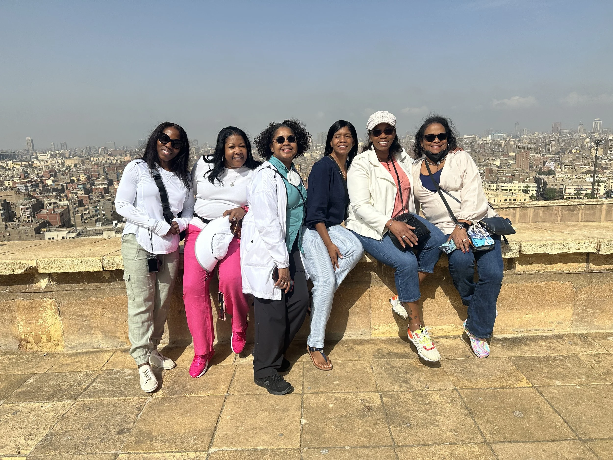 Six women sitting on a stone ledge overlooking a cityscape with tall buildings under a clear sky.