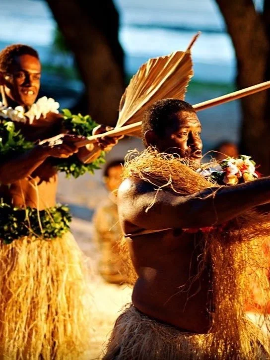 Traditional Polynesian dancers performing with grass skirts and leaf decorations on a beach.