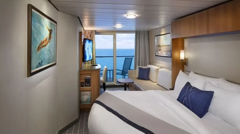 A cruise ship room with a large bed, a sofa with pillows, a small desk with a chair, a balcony with a sliding glass door, and ocean view