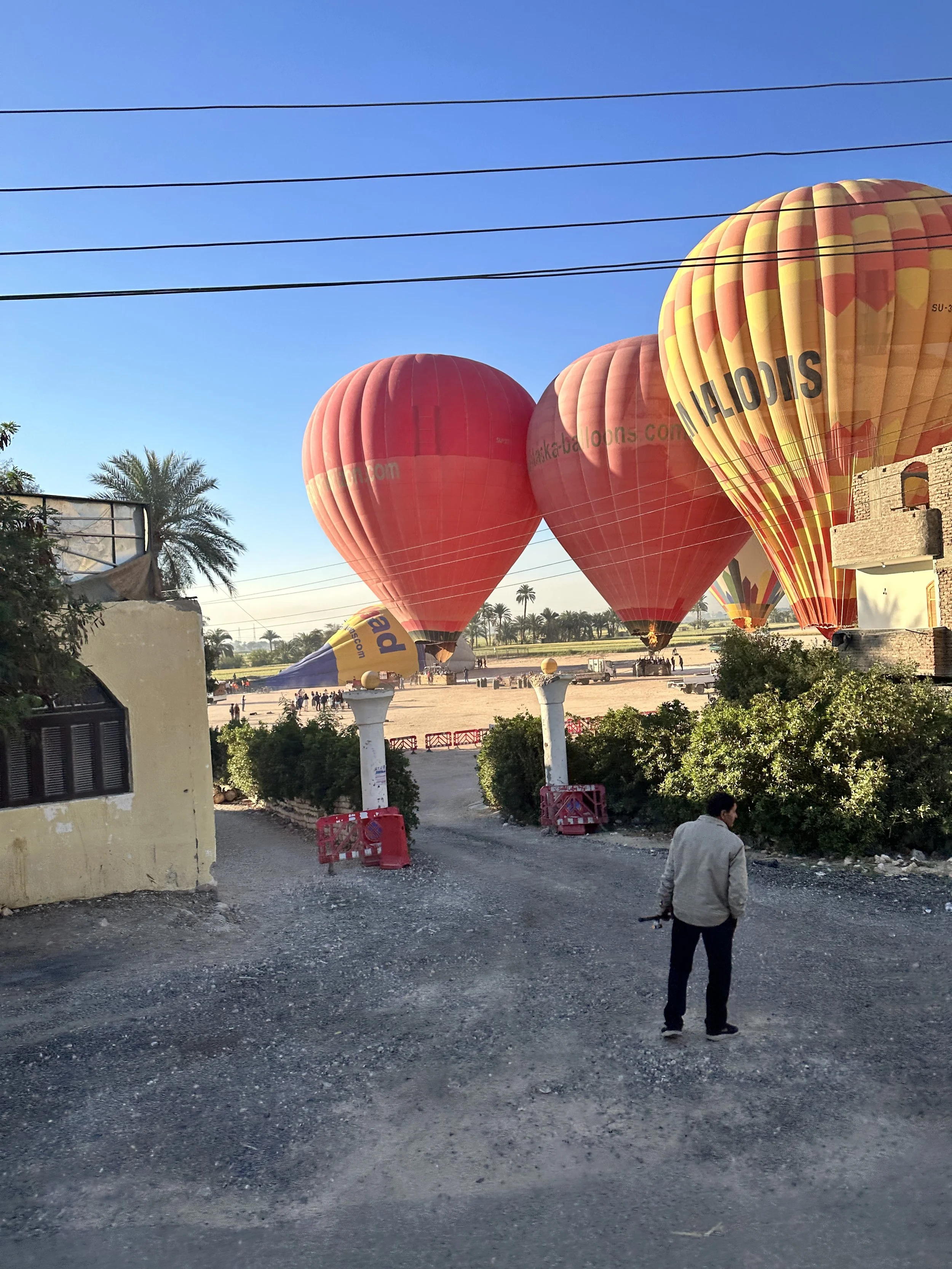 Four hot air balloons being inflated on a dirt lot, with one partially on the ground and the others in the air, under a clear blue sky.