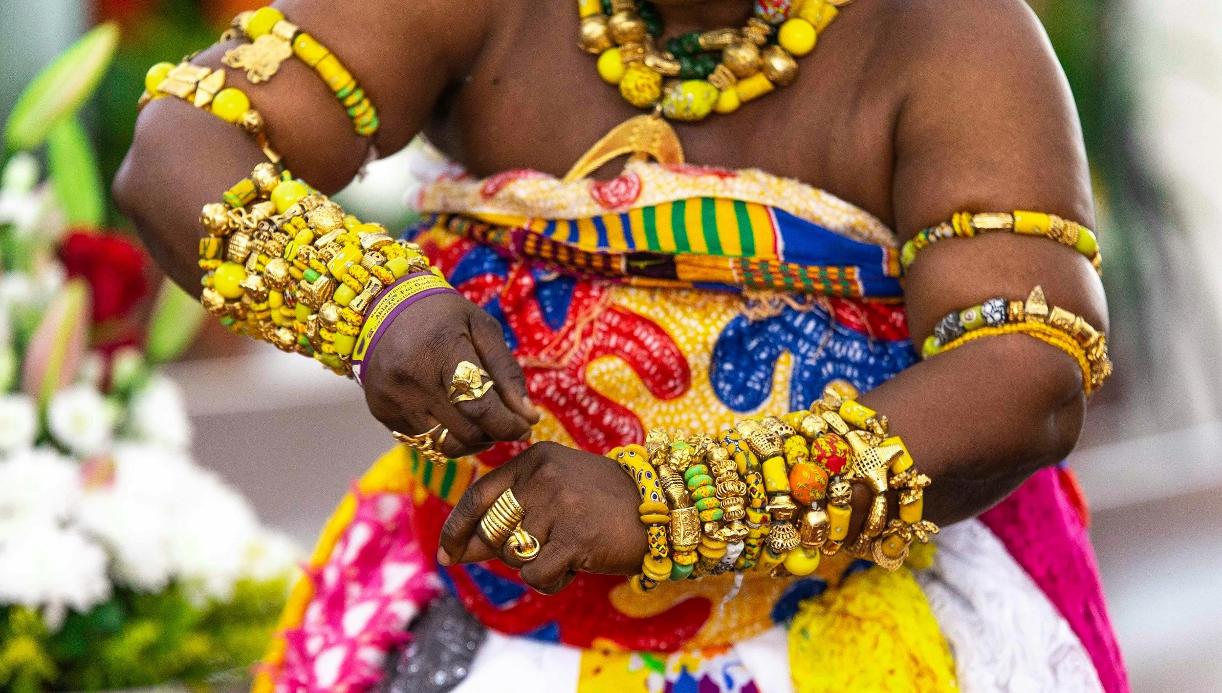 Person wearing colorful traditional clothing with yellow and gold jewelry, including bracelets and rings, and floral background.