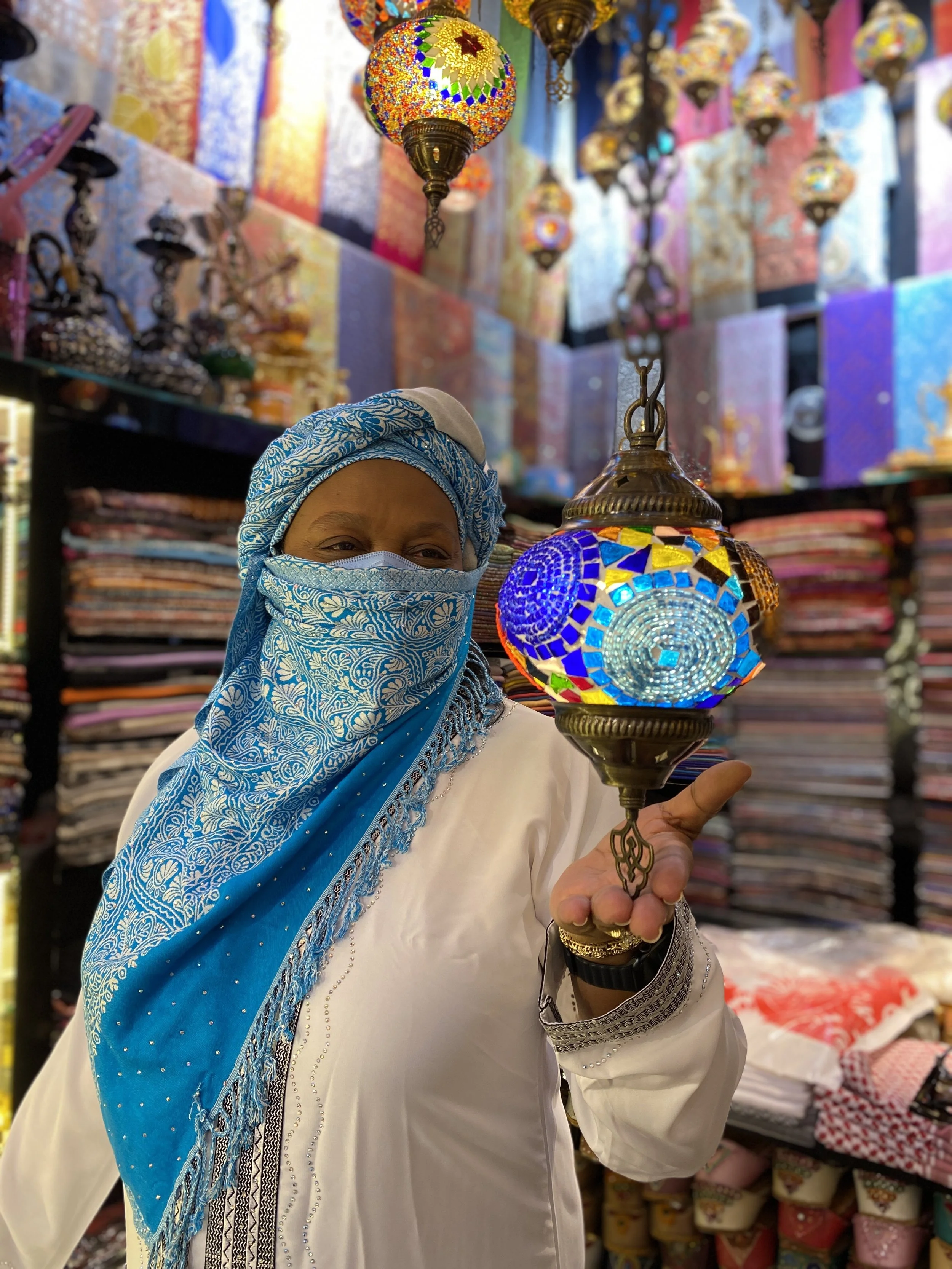 Woman in a white traditional outfit and blue patterned head covering and face mask holding a colorful mosaic Turkish lantern inside a shop with vibrant textiles and other lanterns.