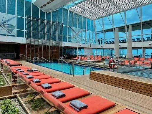 Indoor pool area with lounge chairs and towels, surrounded by large glass windows and a modern architectural structure.