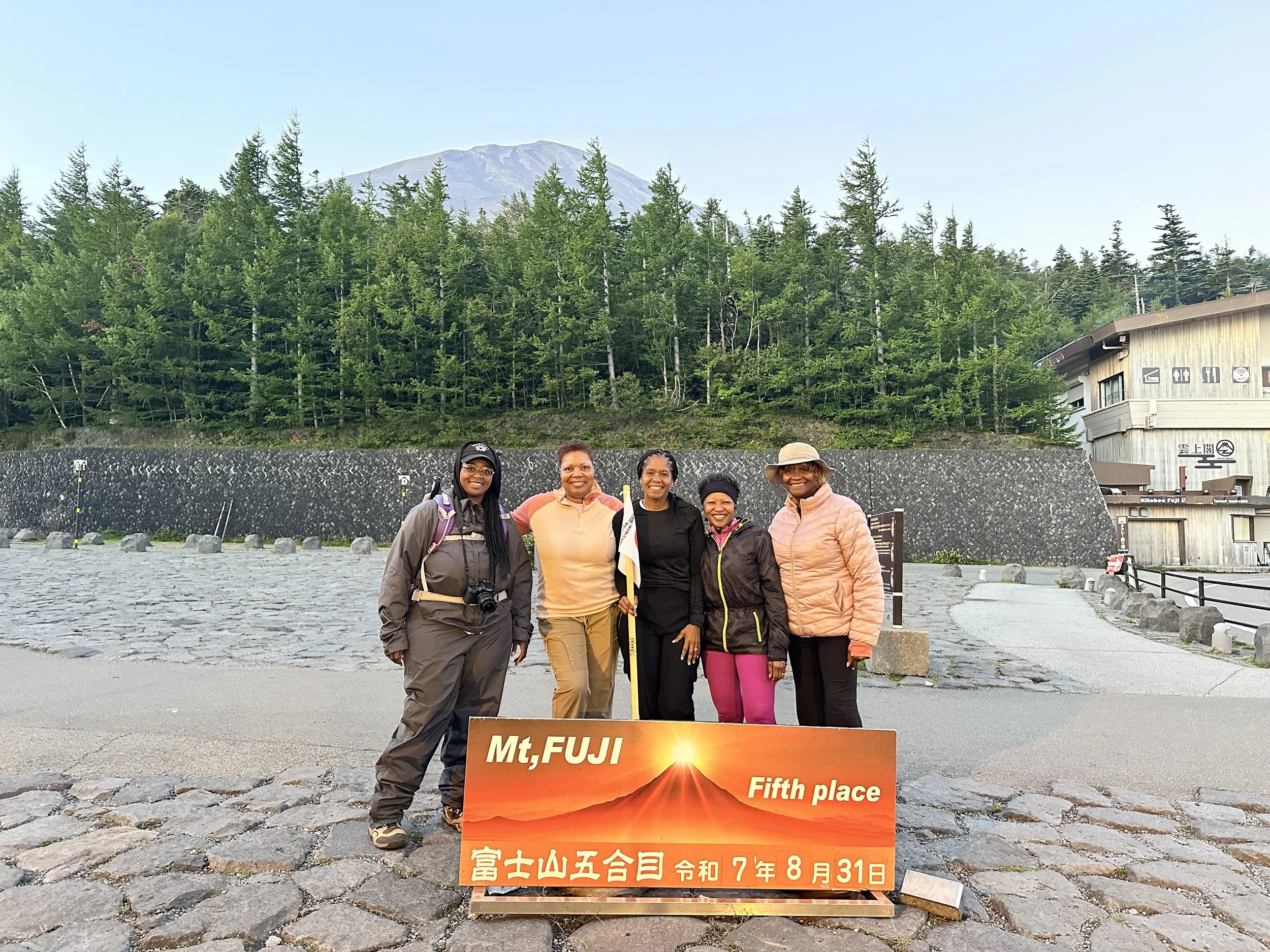 Five women standing together outdoors on a cobblestone pathway, holding a sign that reads 'Mt, FUJI Fifth place' with Japanese characters and a date, in front of a scenic view of Mount Fuji, a forested hillside, and a building.