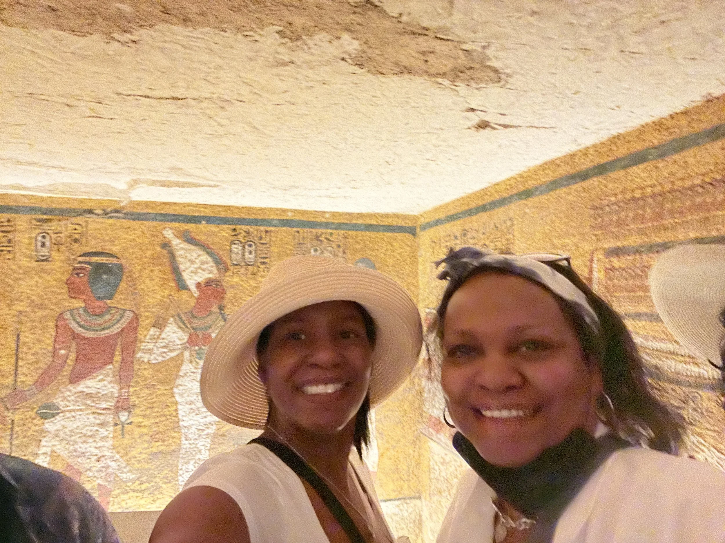 Two women smiling in front of an ancient Egyptian mural with hieroglyphs and figures.