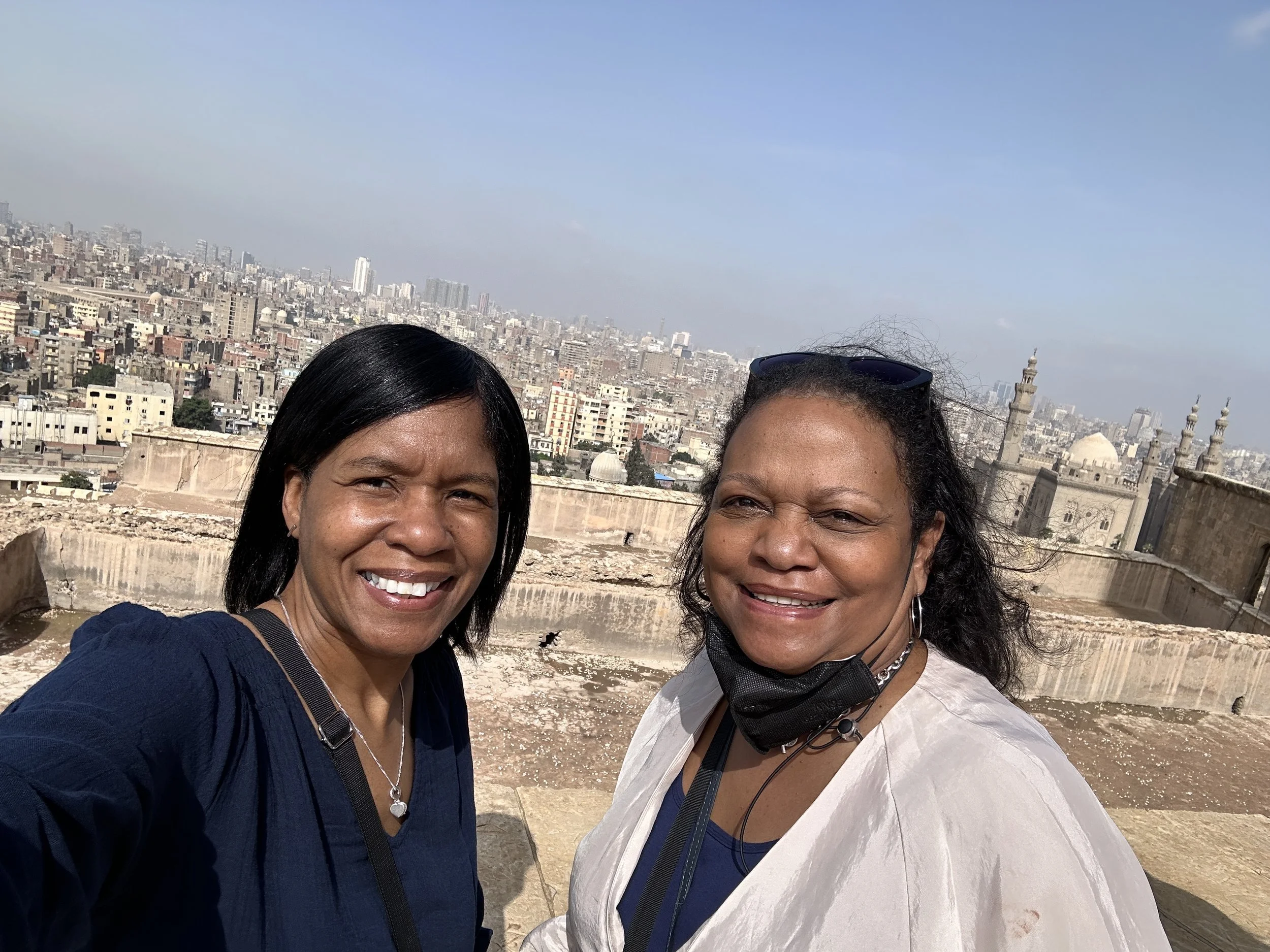Two women smiling for a selfie with a cityscape and a mosque with minarets in the background under a clear sky.