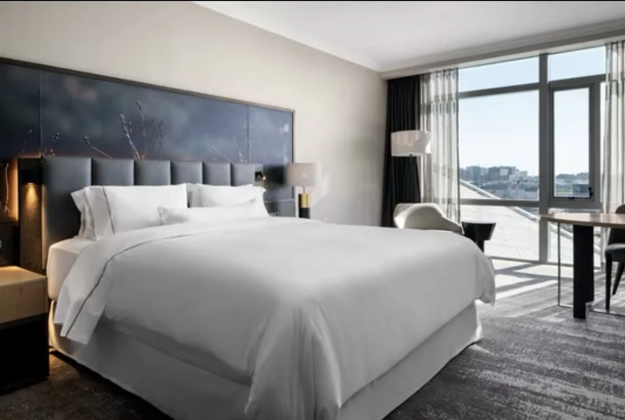 Modern hotel room with a large bed, white bedding, nightstand, lamp, and a balcony with city view.