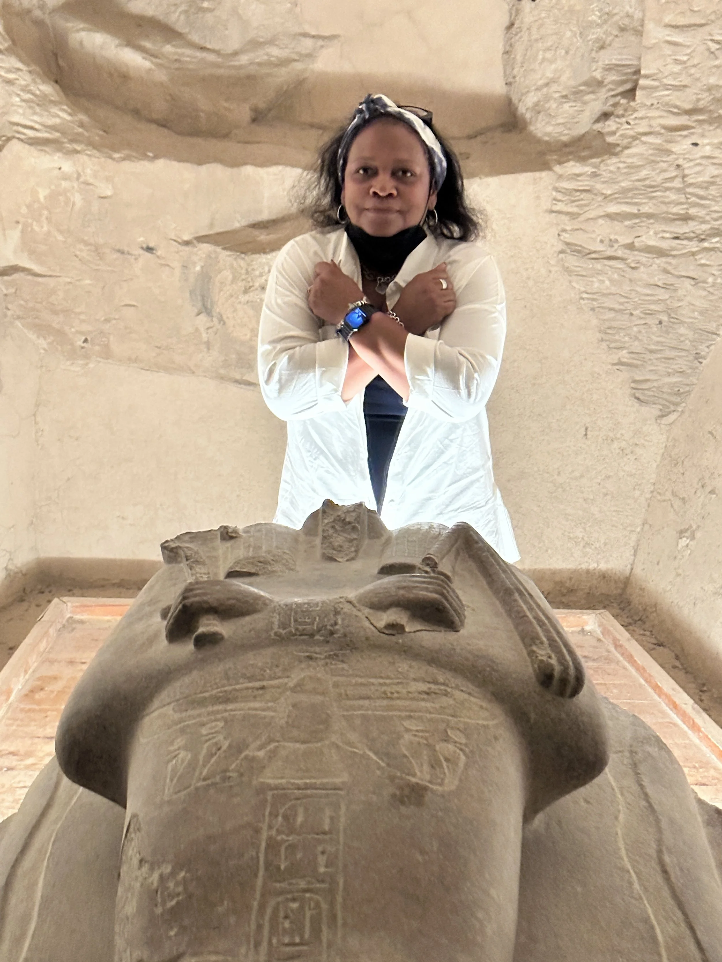 A woman standing behind an ancient Egyptian statue, with her arms crossed over her chest, inside a stone-walled museum or archaeological site.
