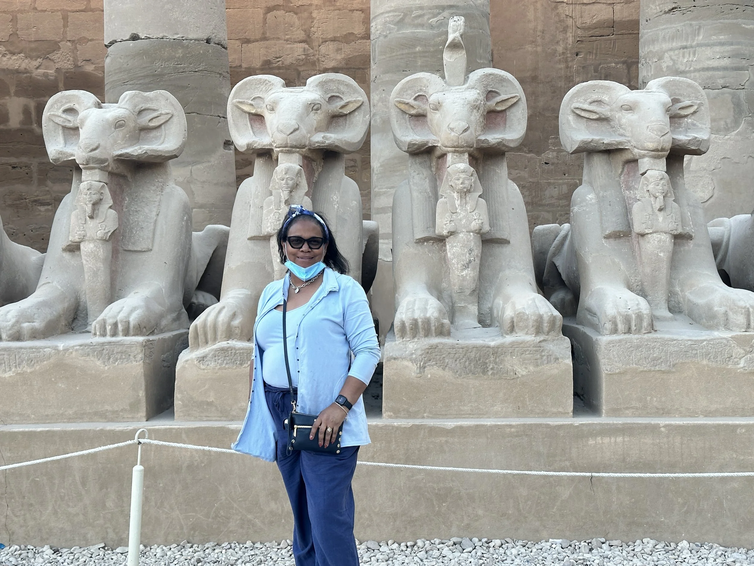 A woman with dark hair, wearing sunglasses, a blue shirt, blue pants, and a blue headband, stands in front of ancient Egyptian statues of sphinxes with ram heads. She has a face mask pulled down under her chin and is smiling at the camera.