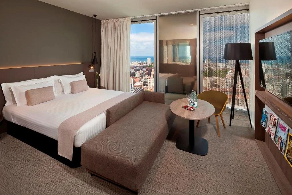 Modern hotel room with bed, sofa, table, and city view through large windows.