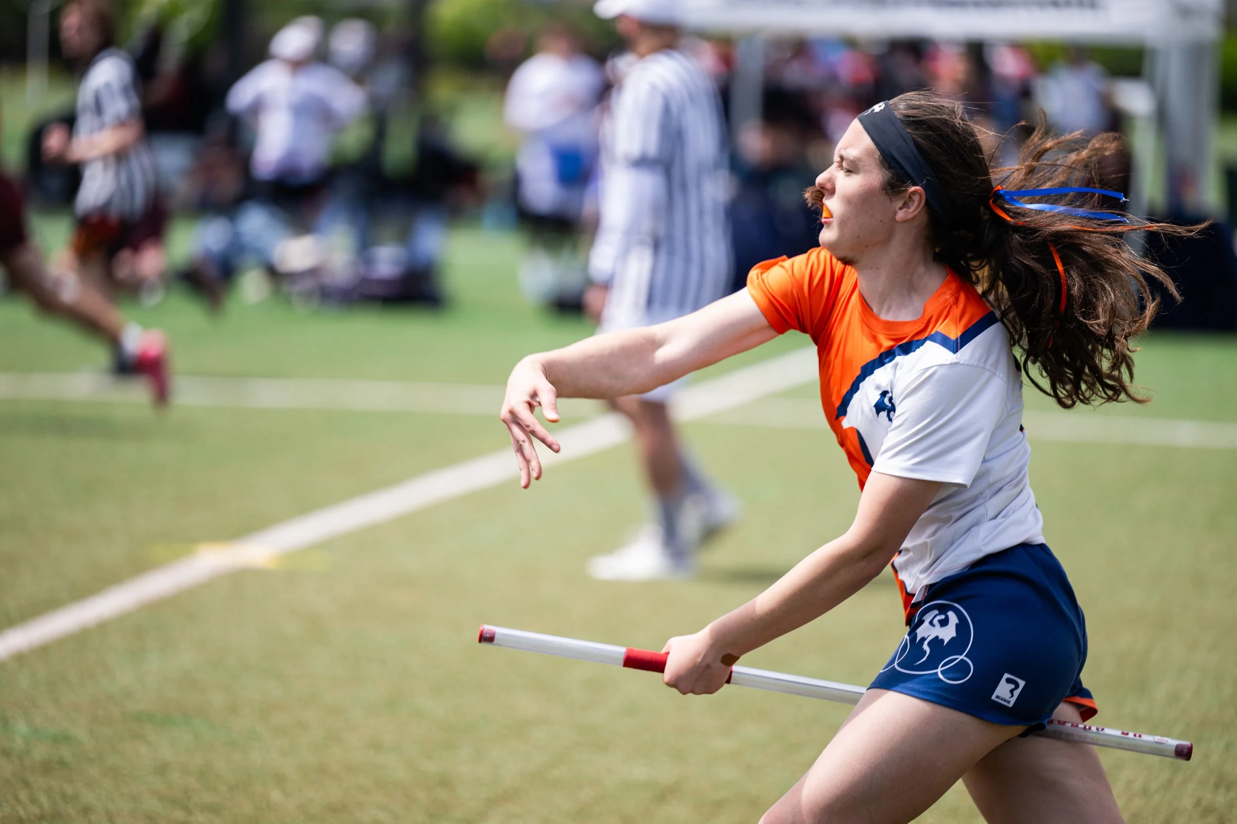 Female field hockey player in orange and white uniform with a black headband, crouching and holding a white and red field hockey stick on a grassy field during a game.