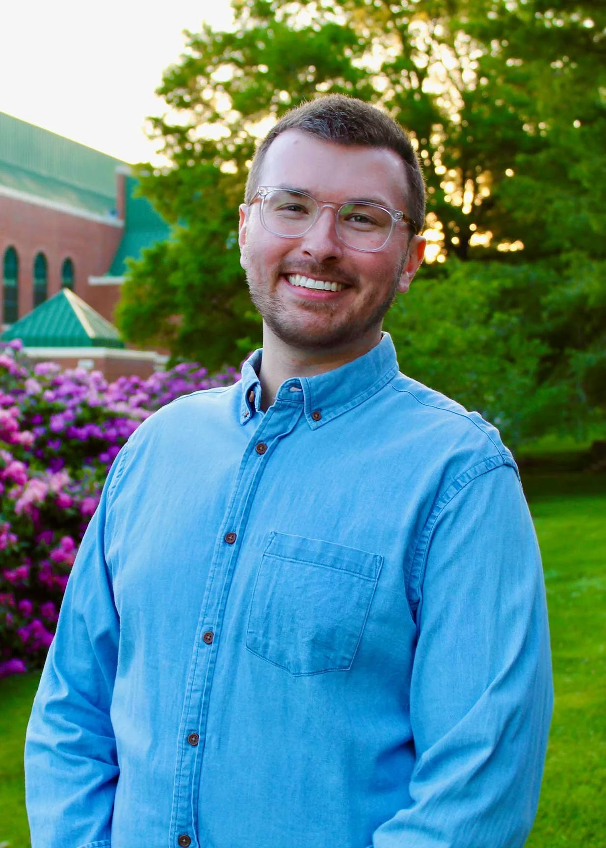 A young man with short brown hair, glasses, and a beard, smiling outdoors during sunset, standing in front of green trees, purple flowers, and a building with a green roof.