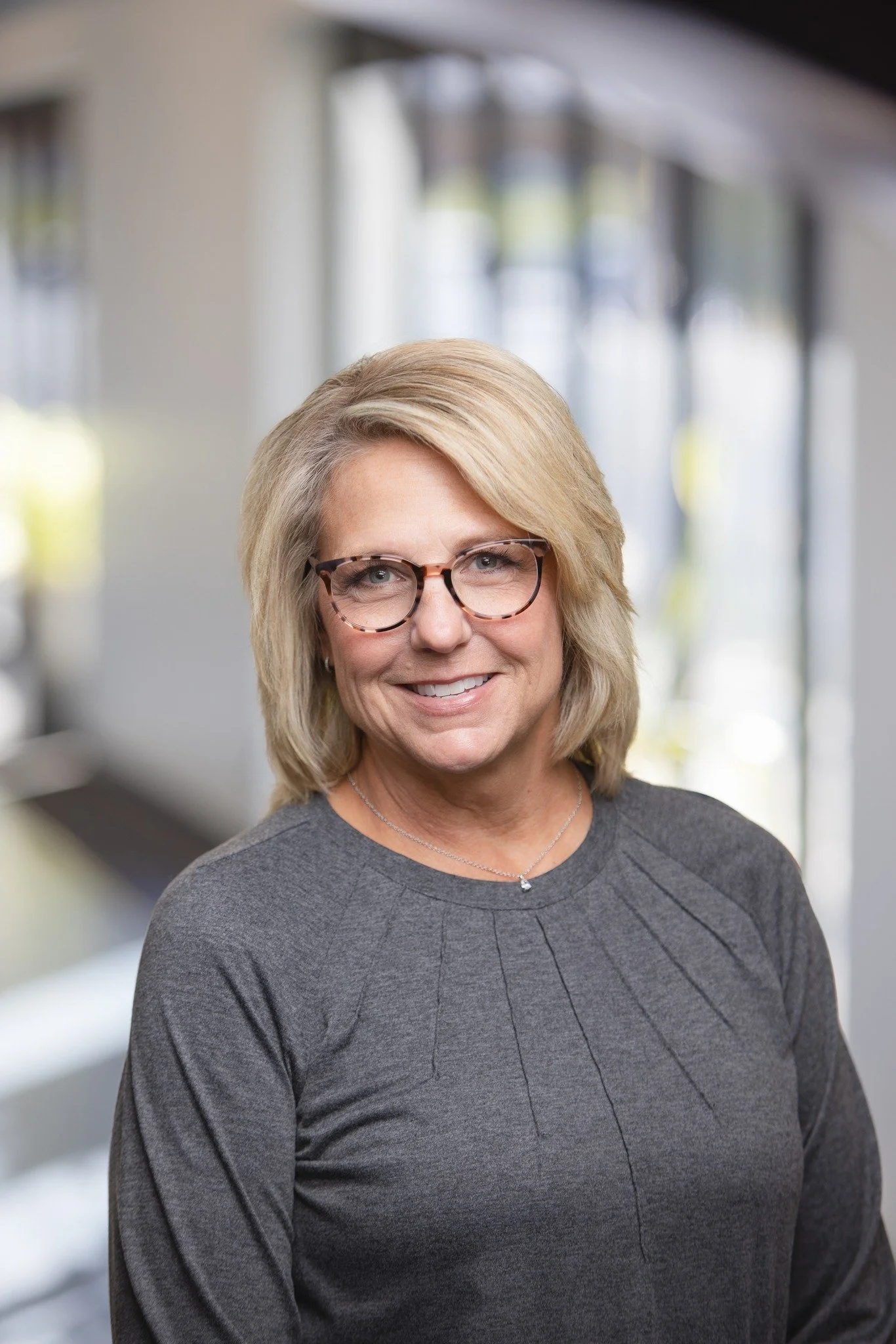 A woman with blonde hair, glasses, and a gray shirt smiling in an indoor setting with bright windows in the background.