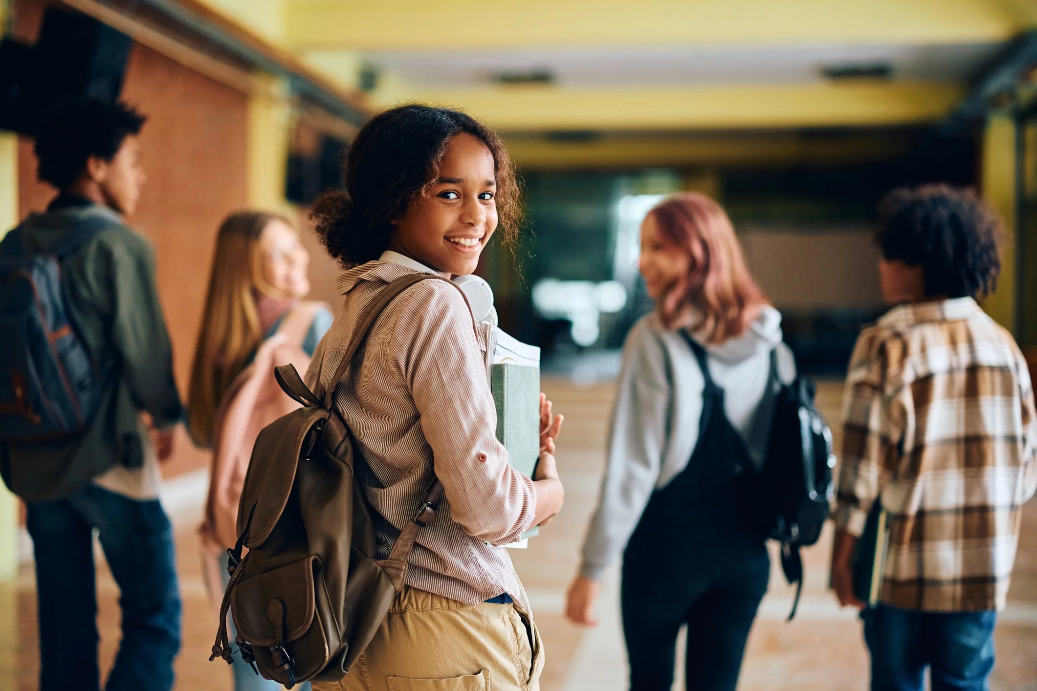 A smiling girl with curly hair holding books and wearing a backpack, standing in a school hallway with other students in the background.
