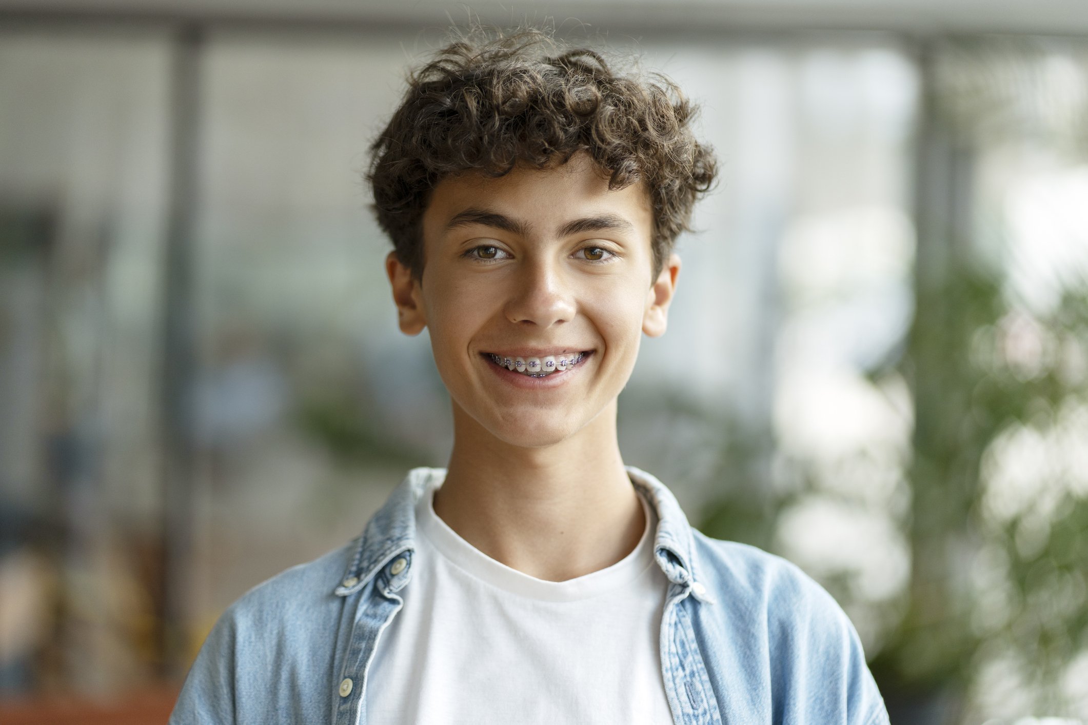 A teenage boy with curly brown hair, braces, and light skin, smiling and wearing a denim jacket over a white t-shirt in an indoor setting with blurred background greenery.