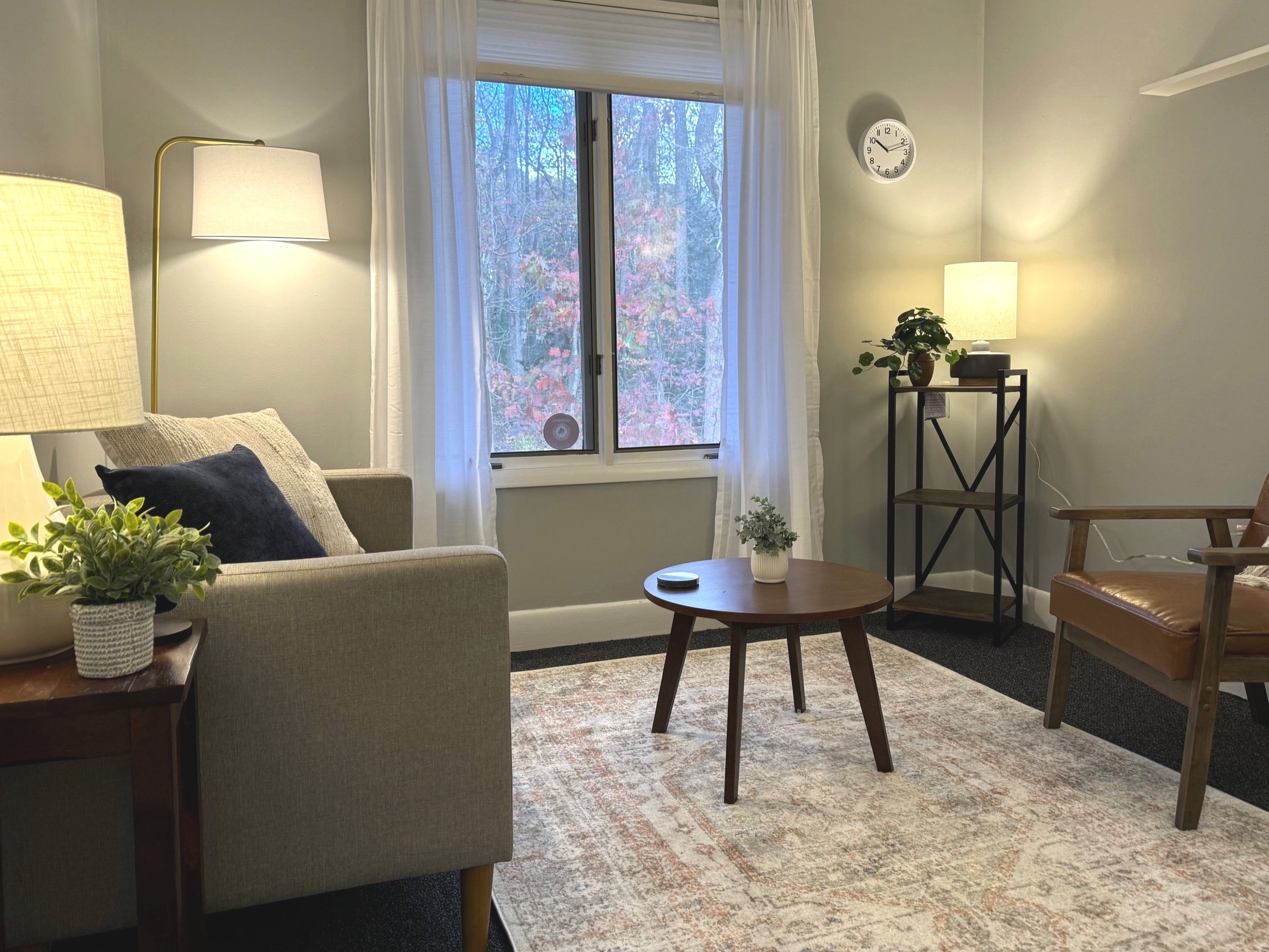 Cozy living room with a grey sofa, brown leather chair, round wooden coffee table, two side lamps, and a small indoor plant. The room features a window with white curtains and a clock on the wall.