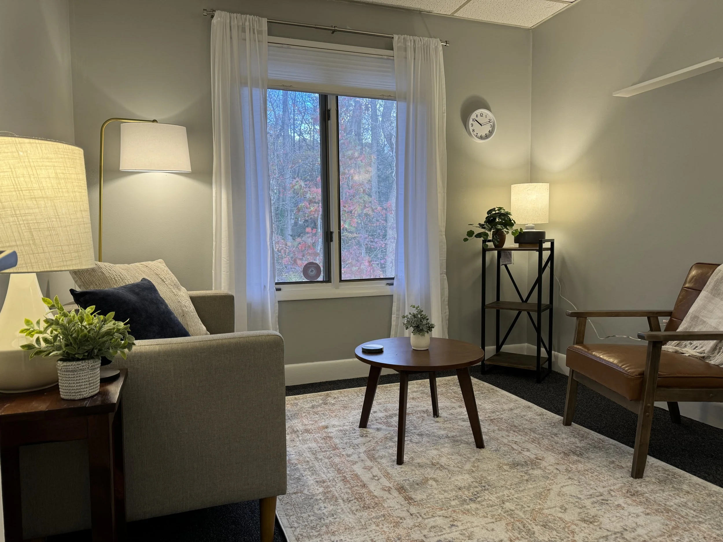 Cozy living room with gray walls, featuring a sofa, armchair, round wooden coffee table, floor lamp, and plants. A window with sheer curtains and two lamps create a warm atmosphere, complemented by a wall clock and a shelf with additional decor.