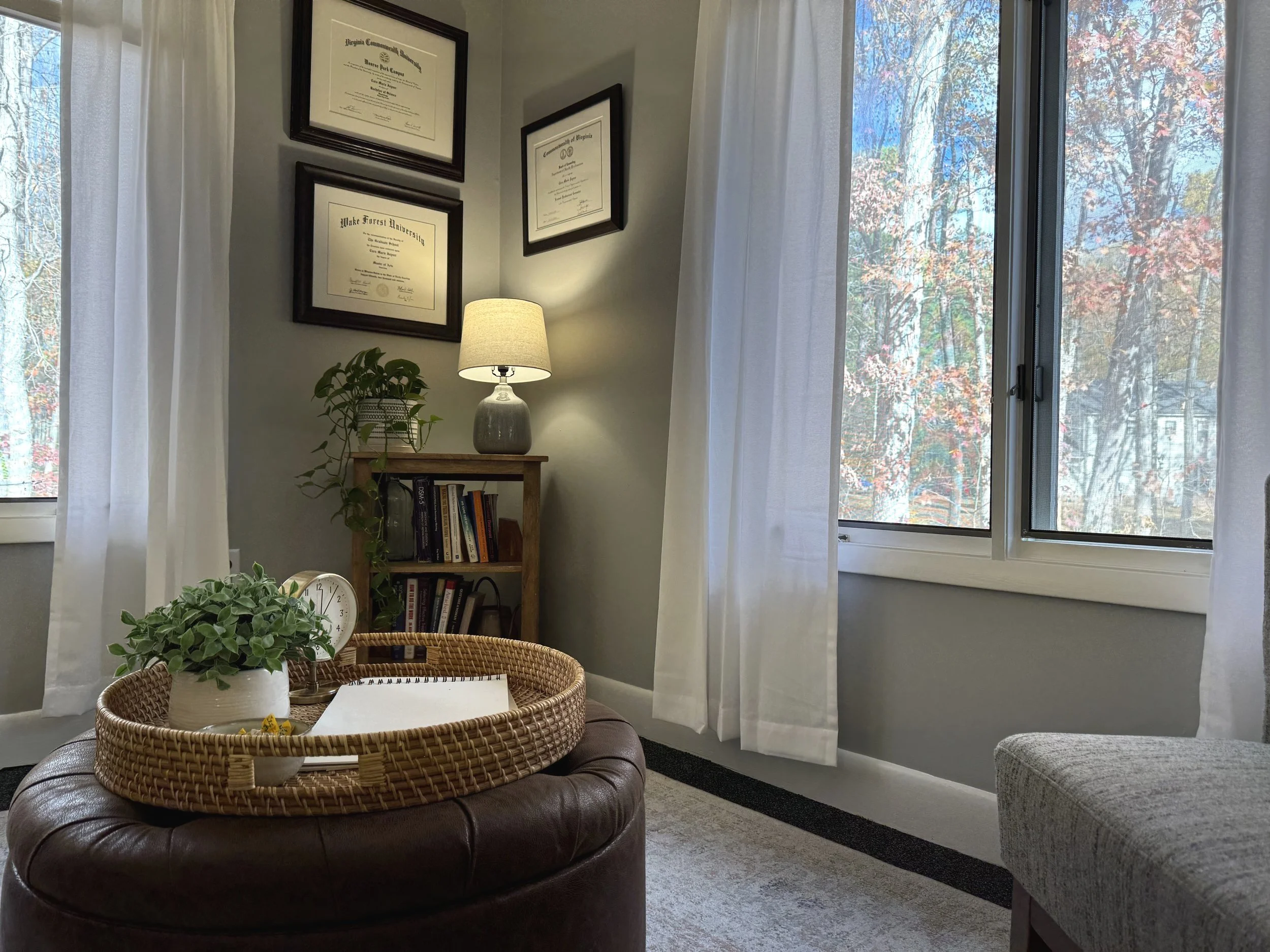 Cozy office corner with diploma certificates on the wall, a wooden shelf with books, a small table with a lamp and plant, a window with white curtains, an ottoman with a woven tray, and a journal. Trees can be seen outside the window.