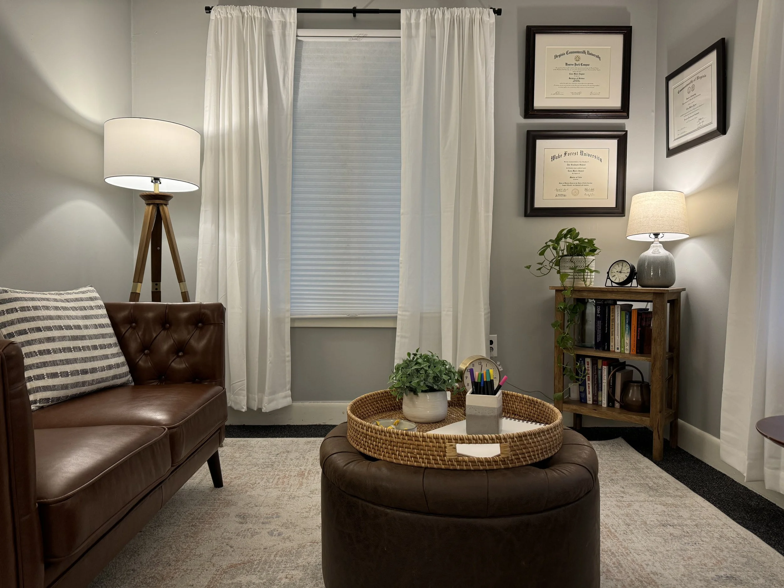 Cozy office or therapy room with a brown leather sofa, floor lamp, window with curtains, framed certificates, bookshelf with books and a plant, rattan tray on an ottoman, and a table lamp on the bookshelf.