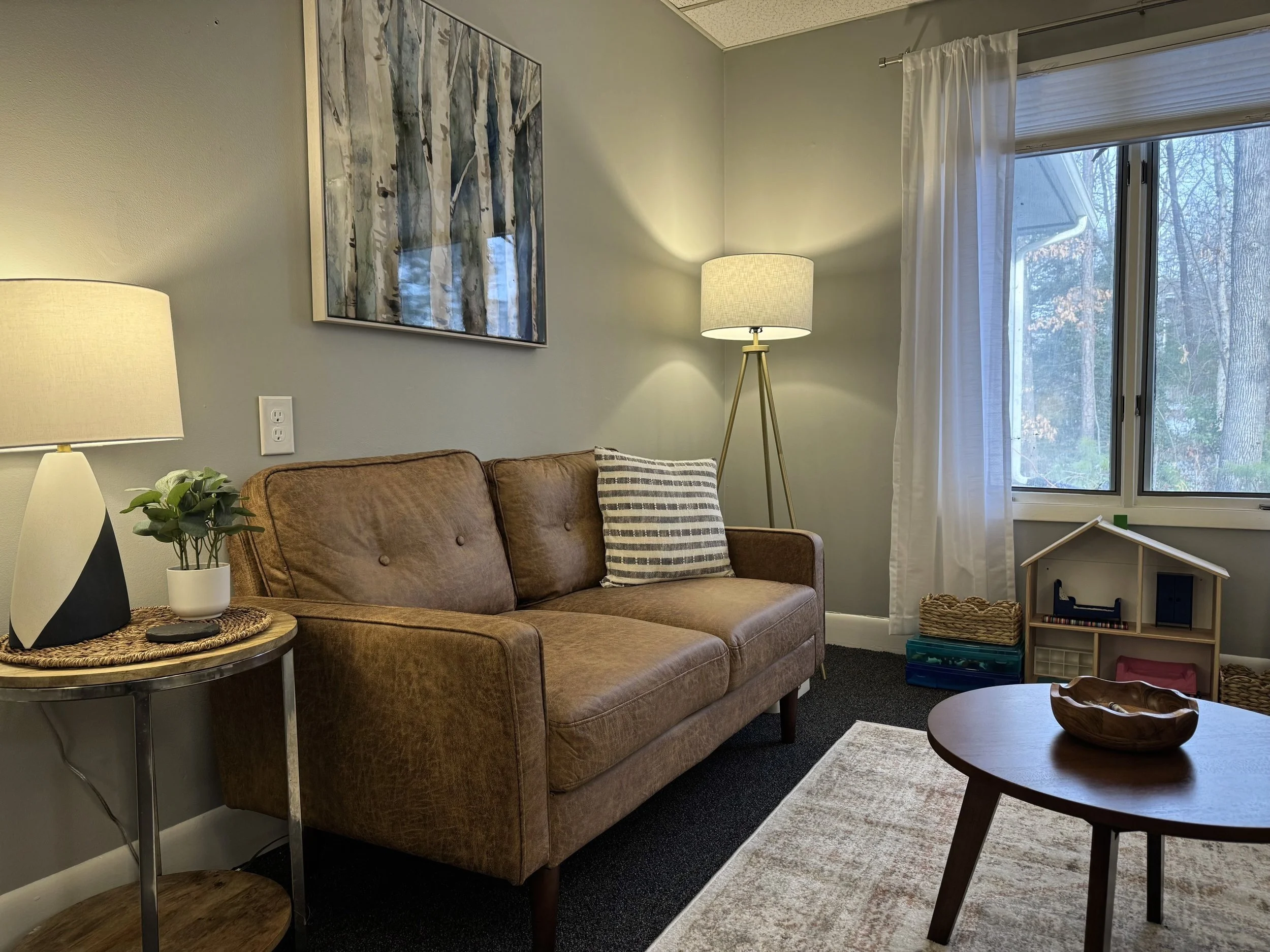 Cozy living room with brown leather sofa, striped cushion, round wooden coffee table, small side table with lamp and plant, floor lamp, window with curtains, wall art, and toy dollhouse with baskets.