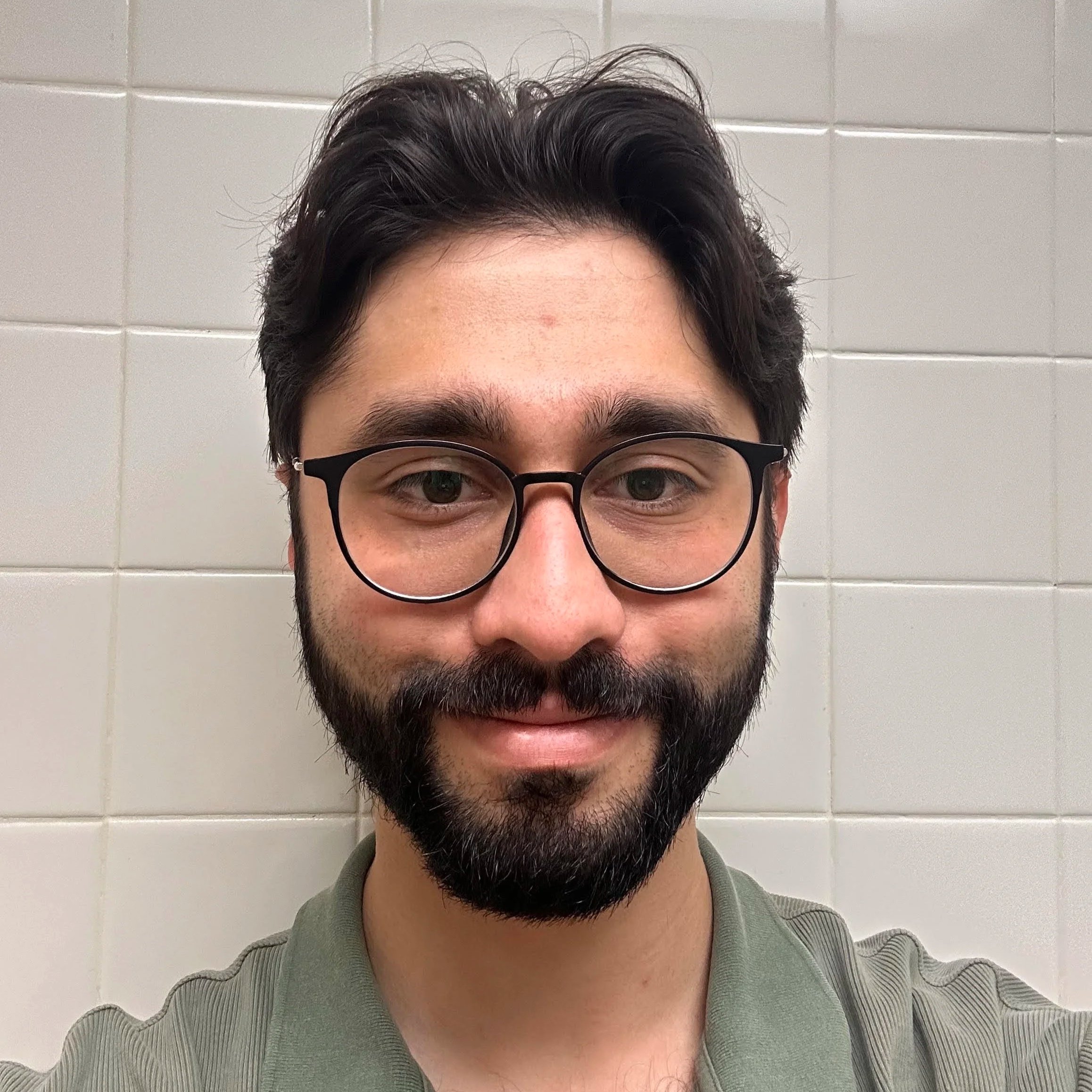 Close-up of a young man with dark hair, glasses, and a beard, smiling in front of a tiled wall.