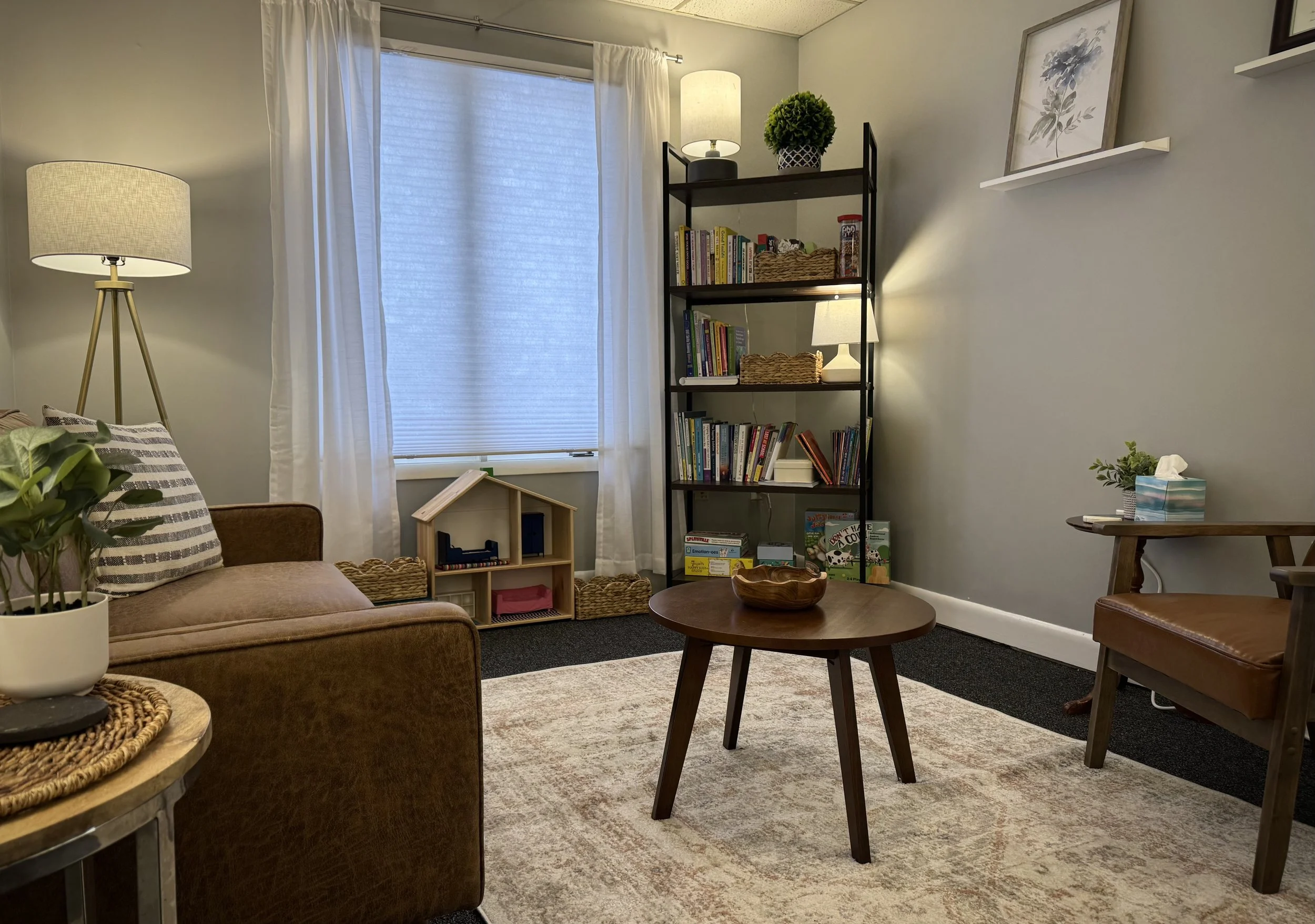 Cozy living room with a brown sofa, wooden coffee table, and tall floor lamp. Bookshelf filled with books and decor items, dollhouse, wicker baskets, and a light-colored area rug. Large window with white curtains, and framed art on the wall.