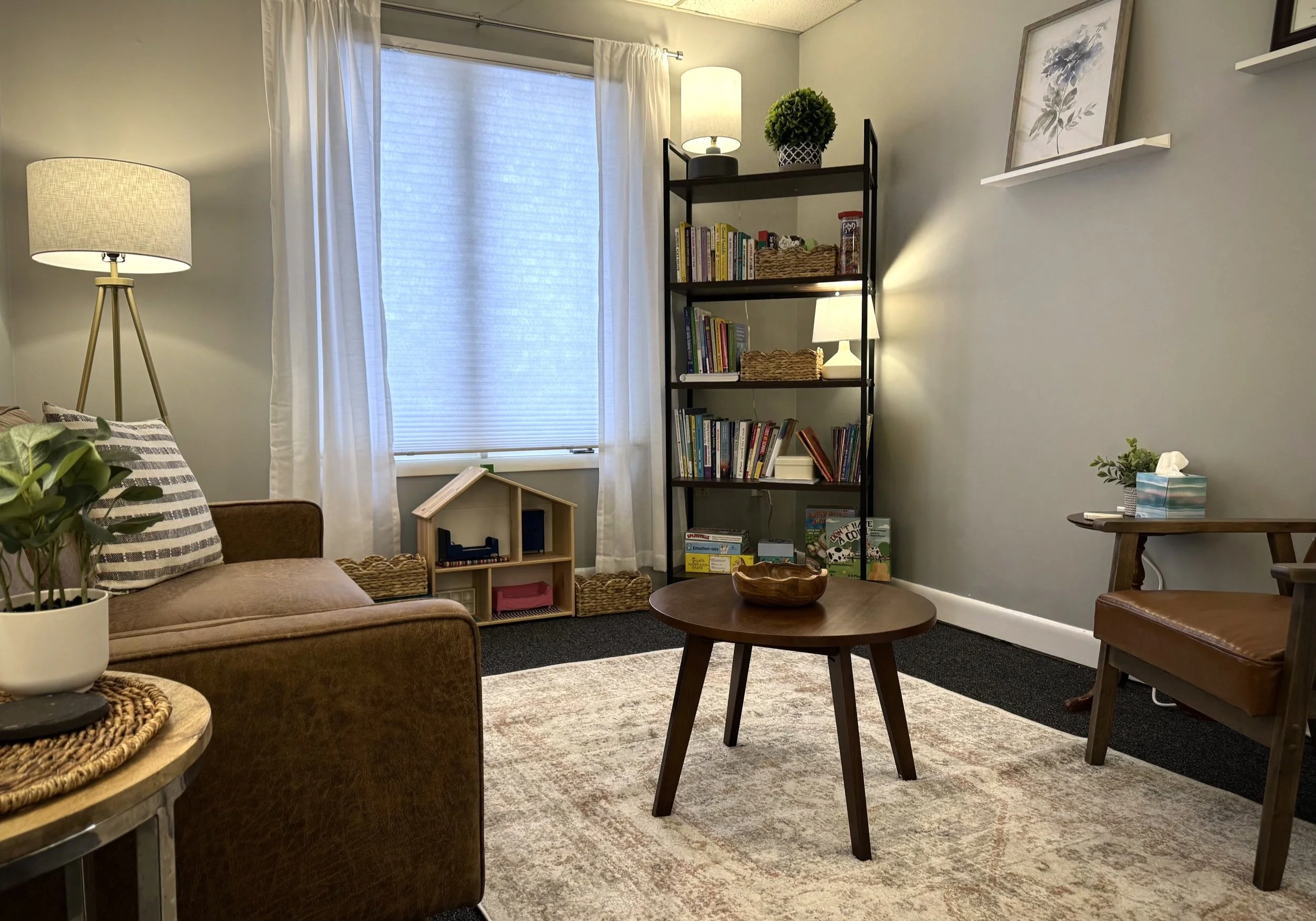 Cozy living room with a brown sofa, wooden coffee table, bookshelf, and small wooden playhouse. Room decor includes a striped pillow, potted plants, and decorative lamps. A window with white curtains is in the background.