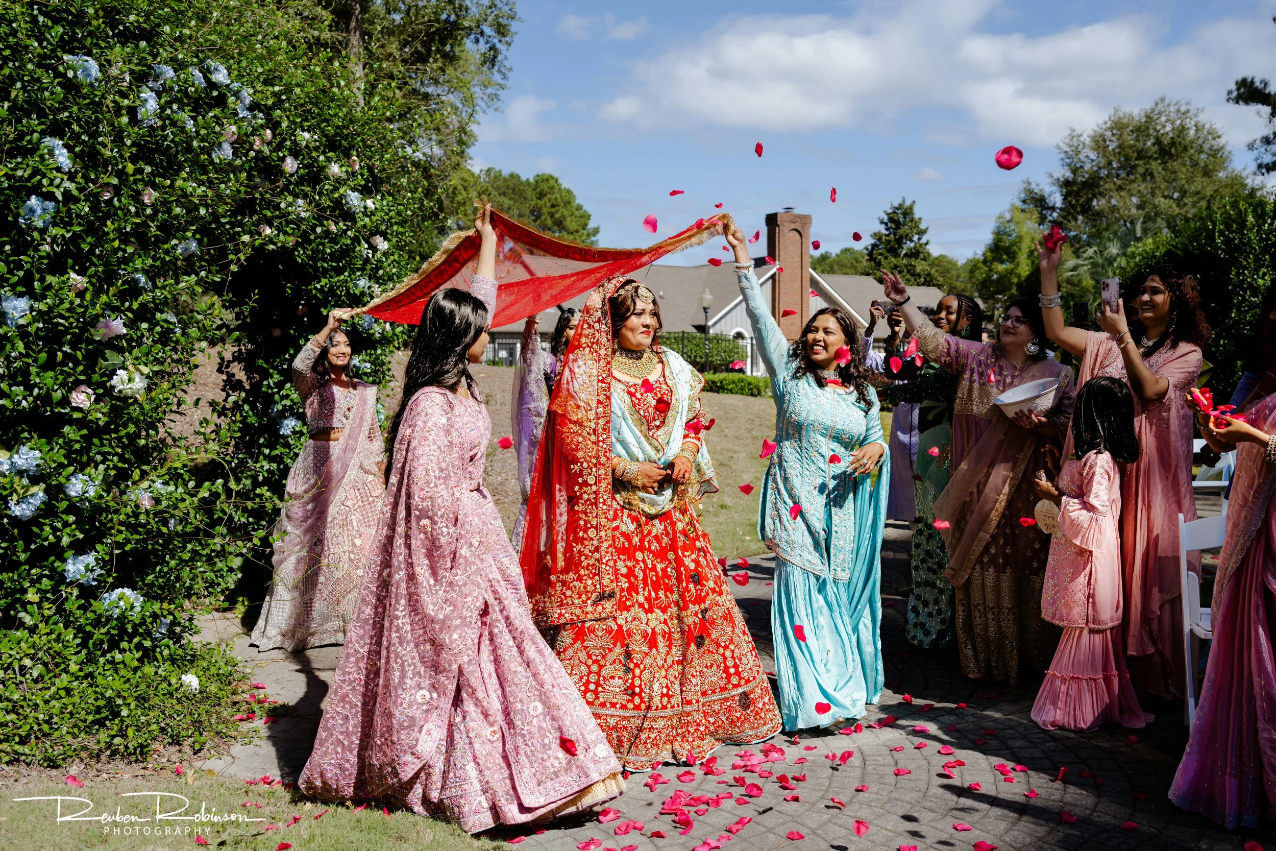 A group of women dressed in colorful traditional South Asian attire at an outdoor wedding celebration, throwing pink rose petals in the air.