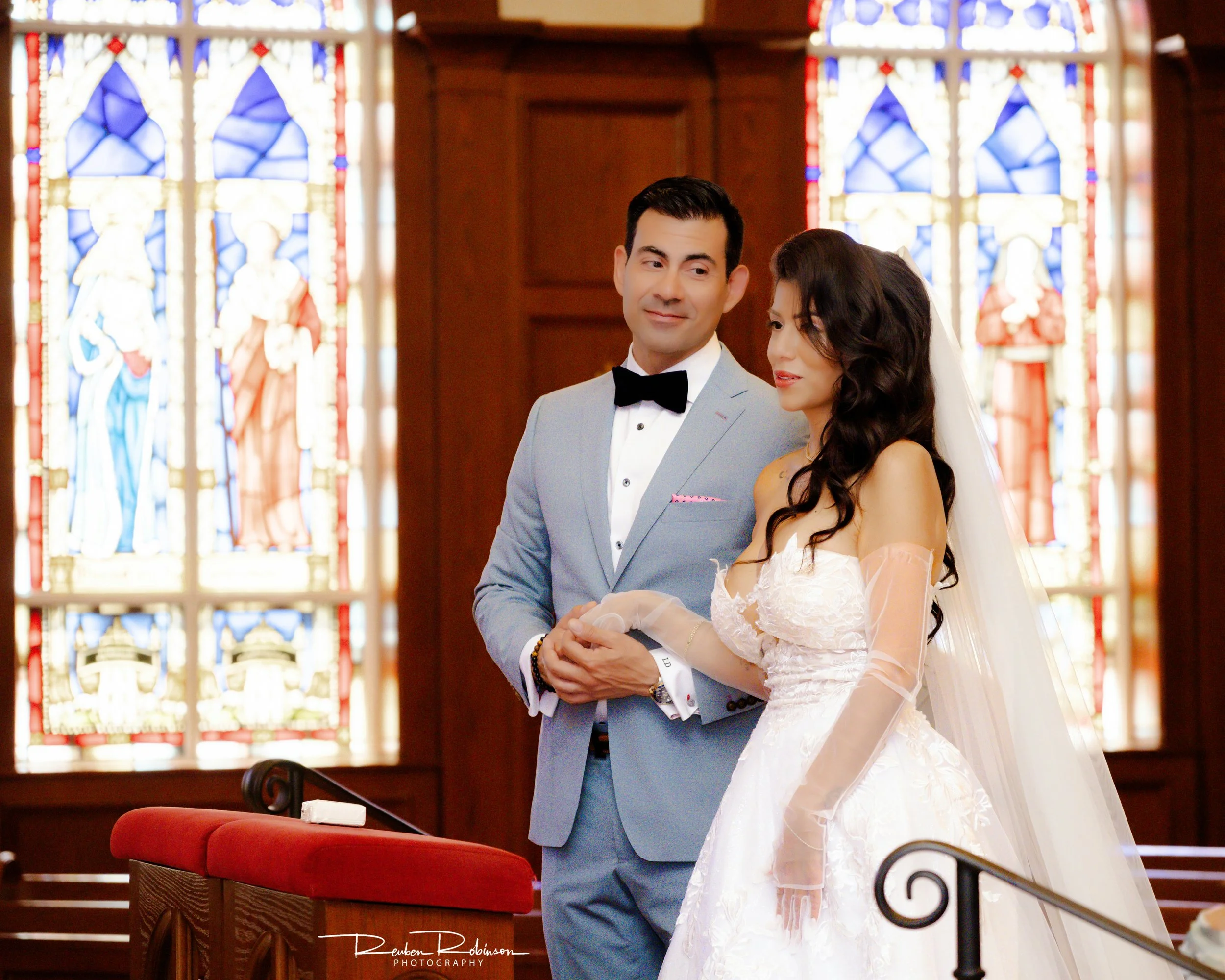 A bride and groom standing inside a church with stained glass windows in the background. The groom is wearing a light gray suit with a black bow tie, and the bride is in a white wedding dress with long dark hair.
