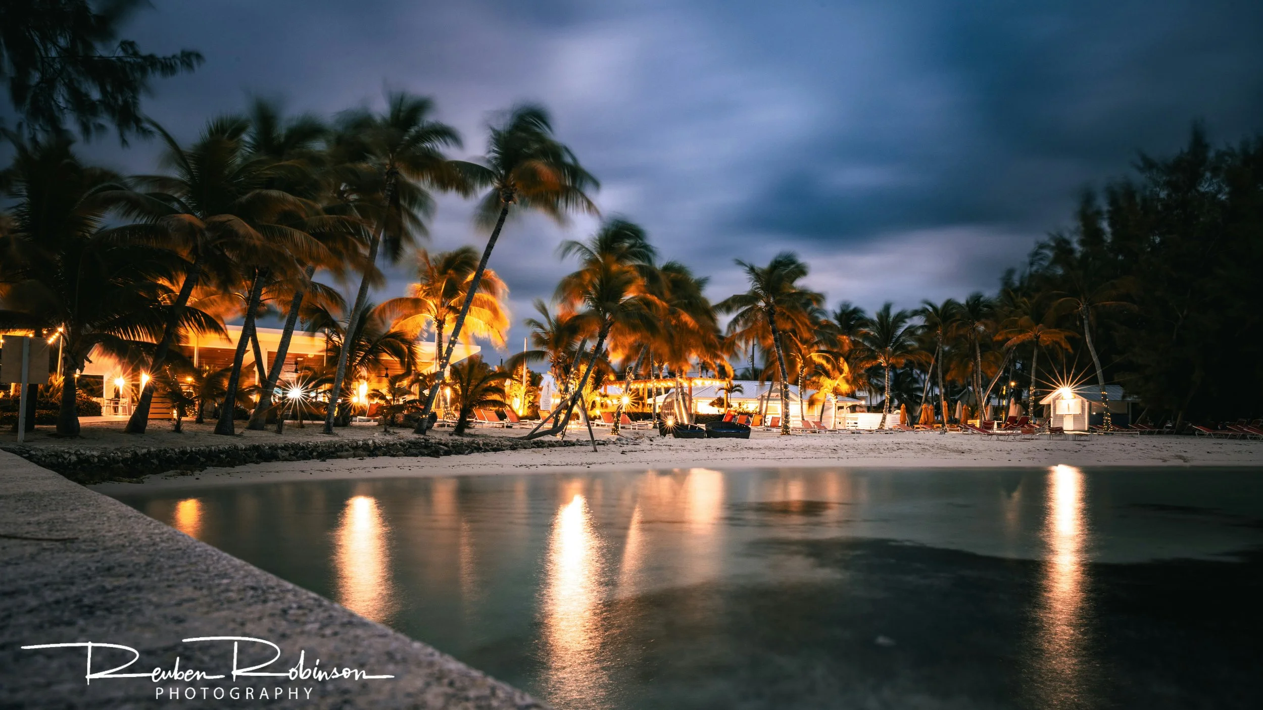Nighttime view of a tropical beach with palm trees, outdoor lights illuminating the area, and a calm body of water reflecting the lights and trees.