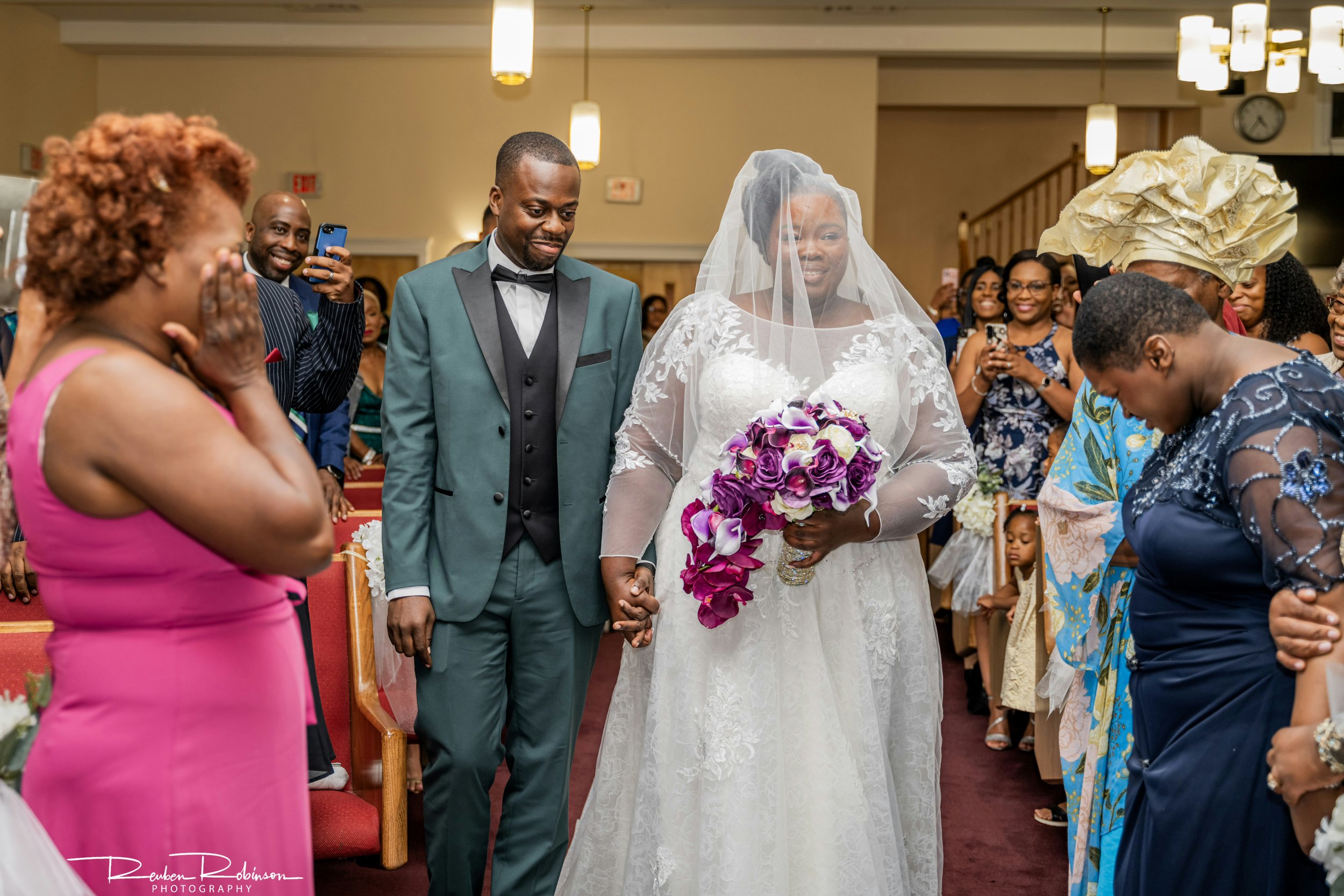 Bride and groom walking down the aisle holding hands, surrounded by smiling and emotional guests, with the bride wearing a white lace wedding dress and veil, holding a bouquet of purple flowers.