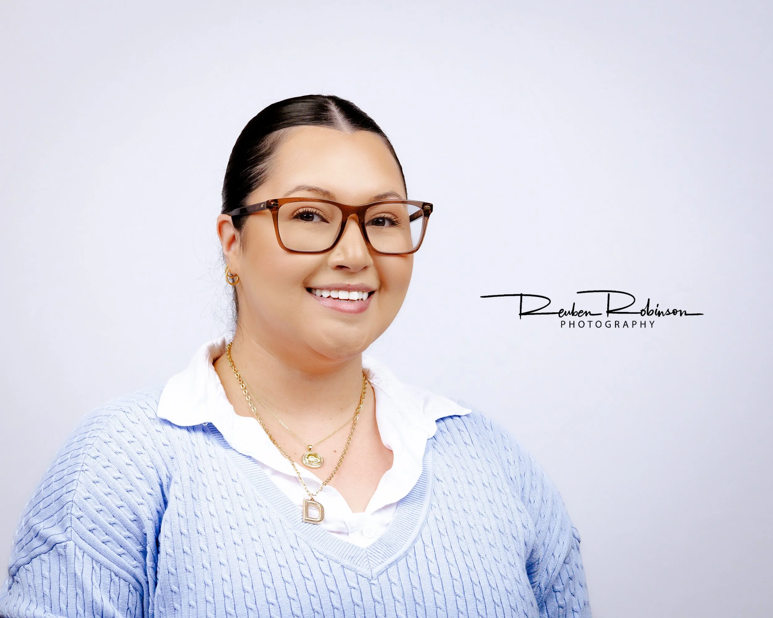A woman with dark hair and glasses smiling, wearing layered necklaces and gold jewelry, against a white background with text 'Reuben Robinson Photography' on the bottom right.