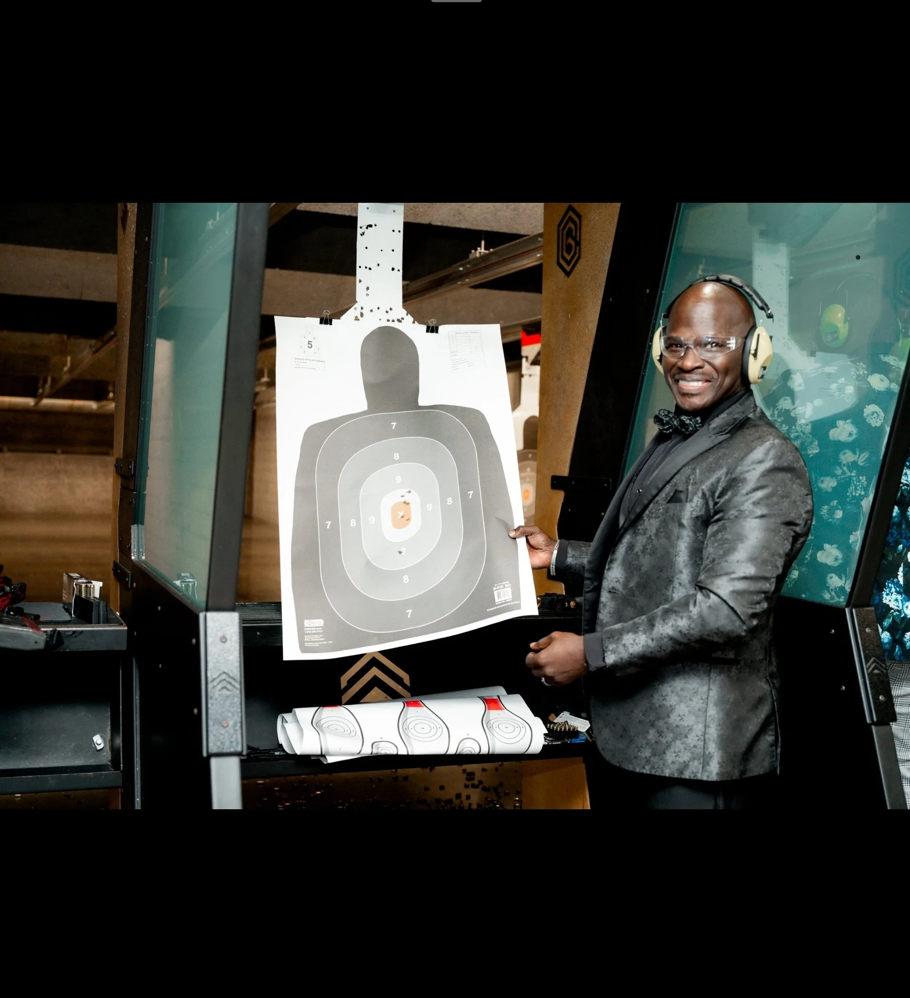 A man in a tuxedo and glasses, wearing hearing protection, is smiling and holding a target paper at a shooting range. The target displays a silhouette with concentric scoring rings and a bullseye.